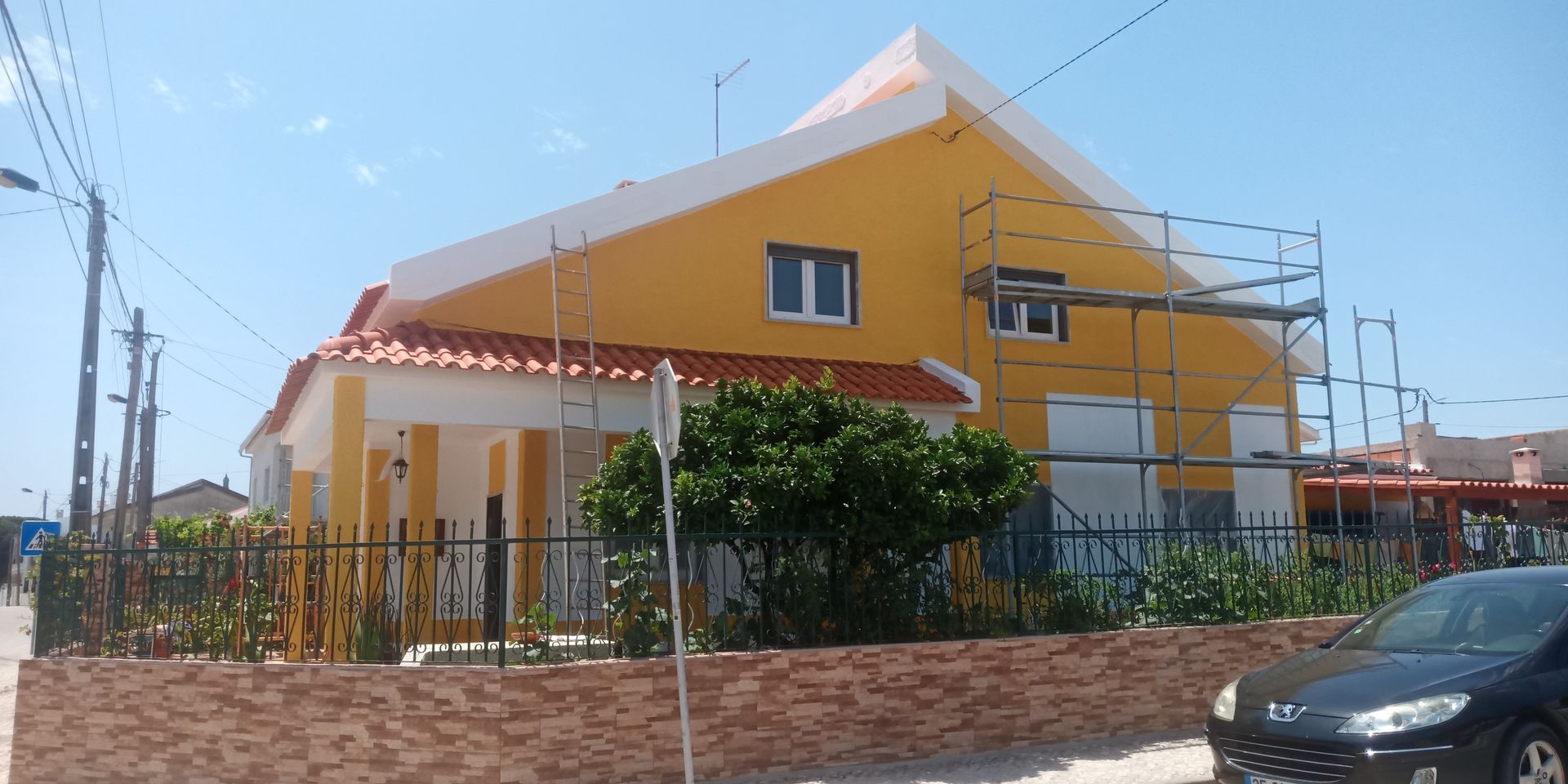 Casa amarela com detalhes brancos, telhado laranja e andaimes. Um carro está estacionado em frente.