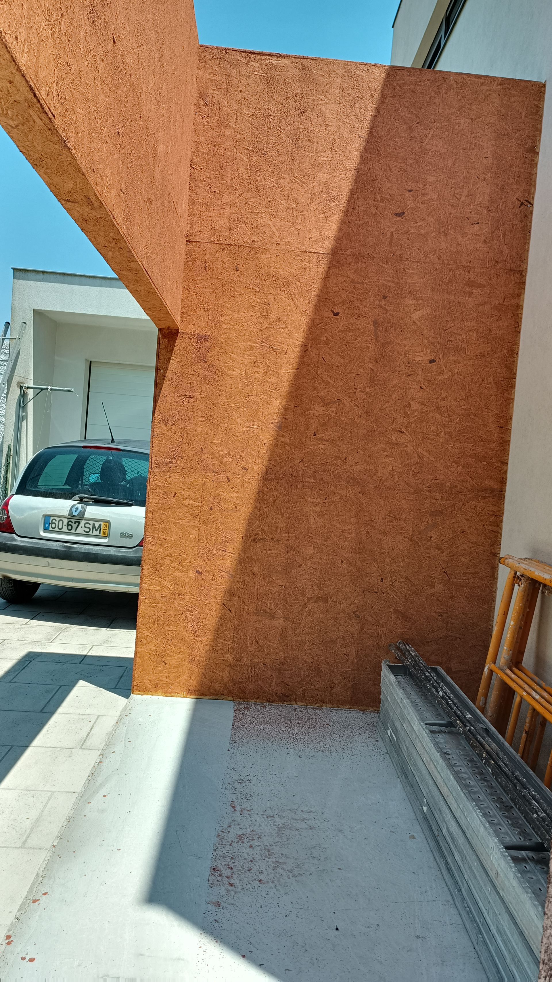 Fachada marrom, semelhante à cortiça, com sombra, ao lado de um carro branco e uma porta.