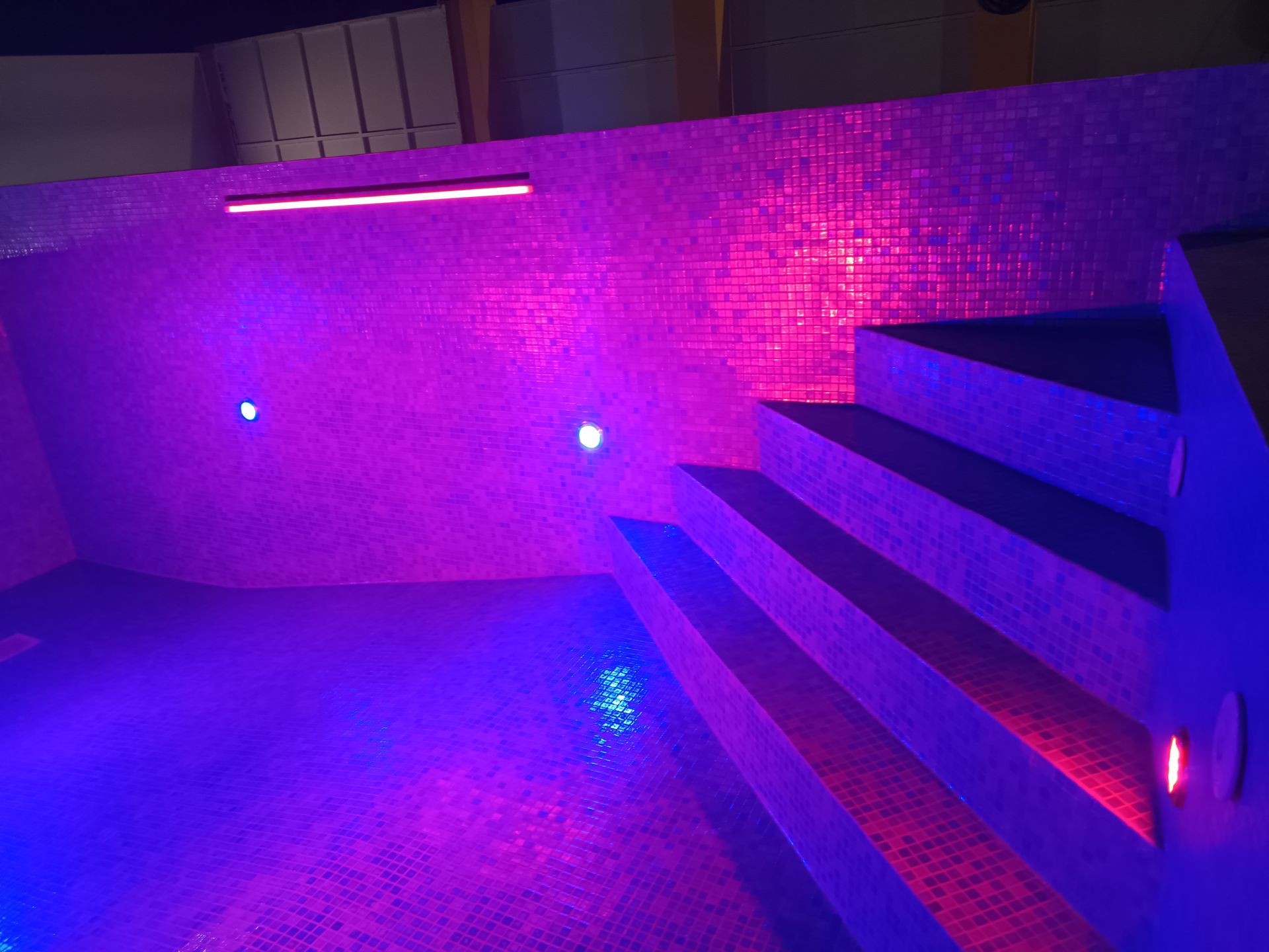 Piscina interna com iluminação roxa, paredes e degraus revestidos com azulejos. Luzes de teto e de parede iluminam a água e os degraus.