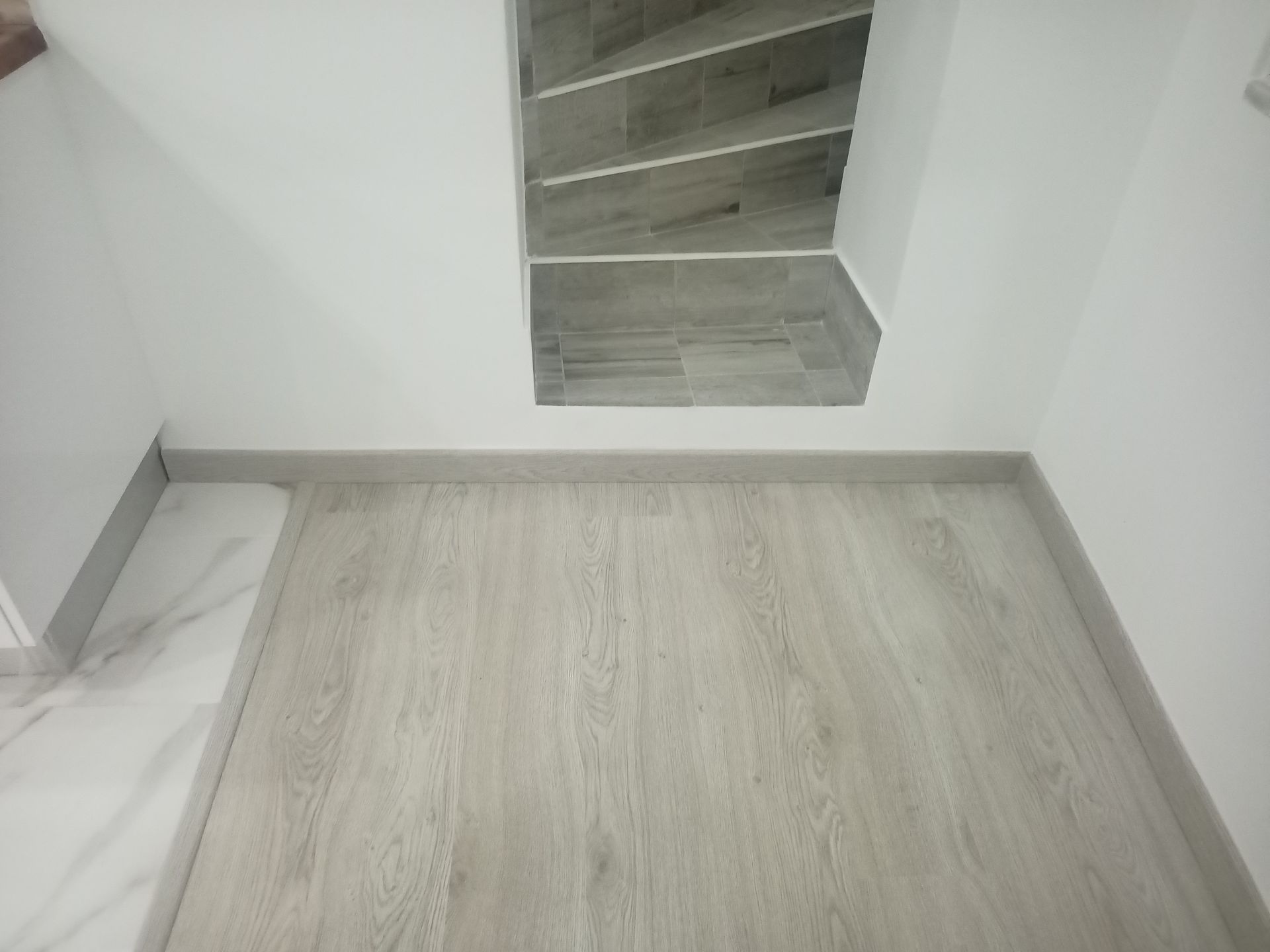 Piso com aparência de madeira clara, paredes brancas e escadas com azulejos cinza.