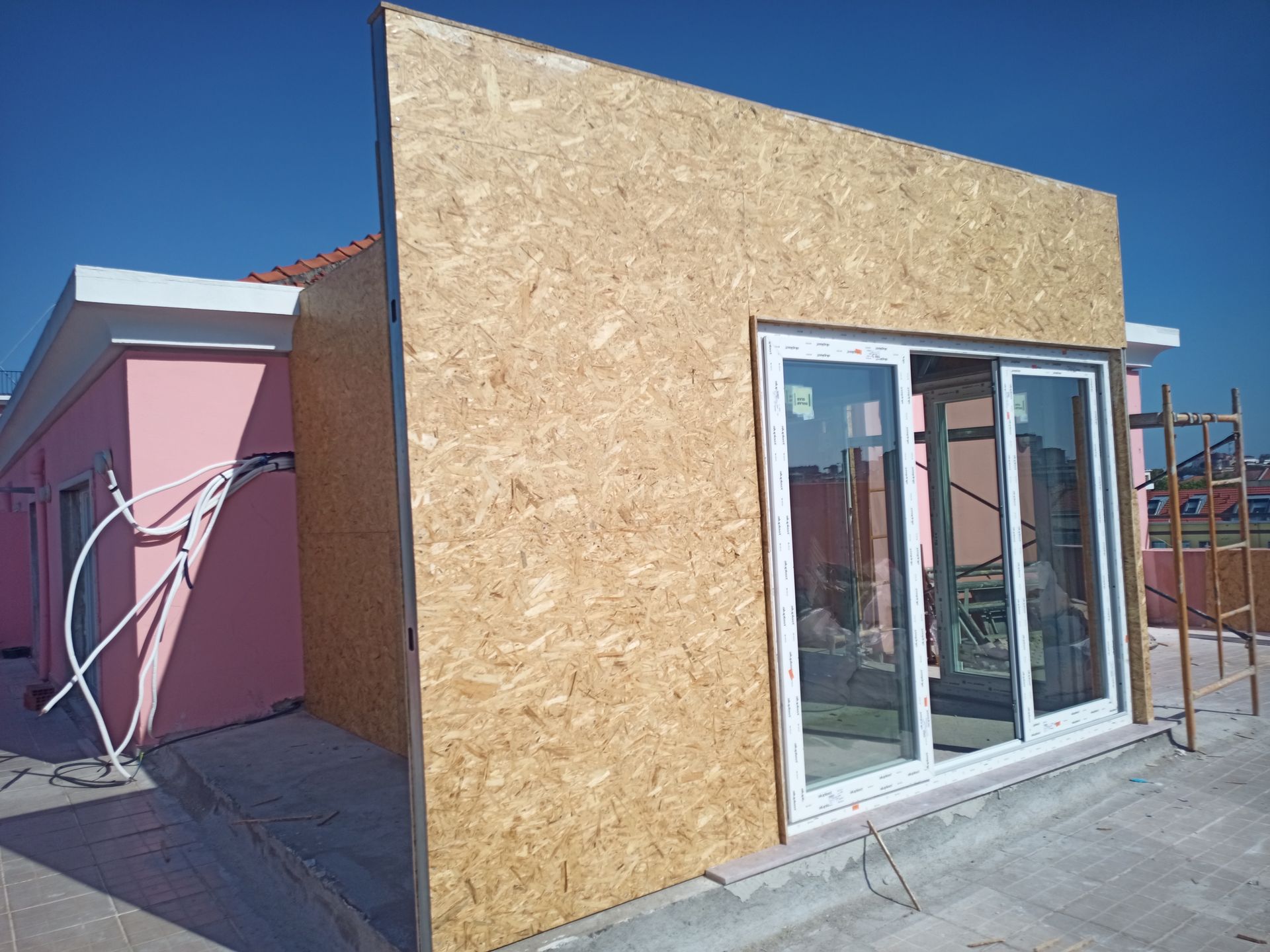 Canteiro de obras com parede revestida de OSB, porta de vidro deslizante e um prédio rosa sob um céu azul.