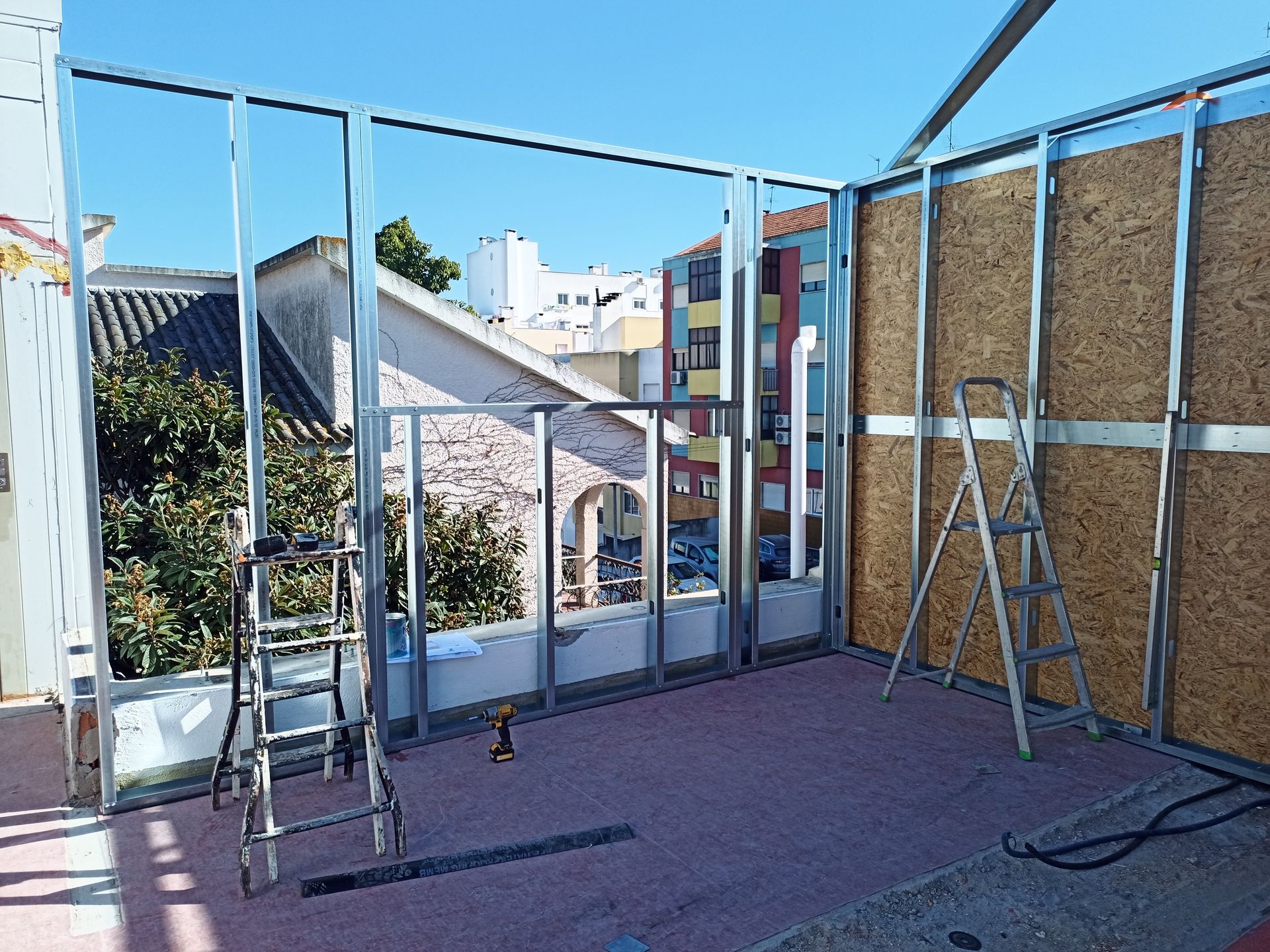 Construção de um cômodo com estrutura metálica, madeira compensada e vista para outros prédios. Duas escadas presentes.