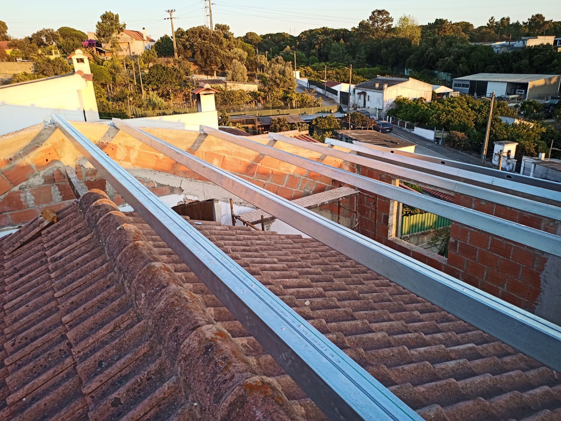 Telhado com vigas metálicas sendo instaladas, cercado por telhas vermelhas em um ambiente suburbano.