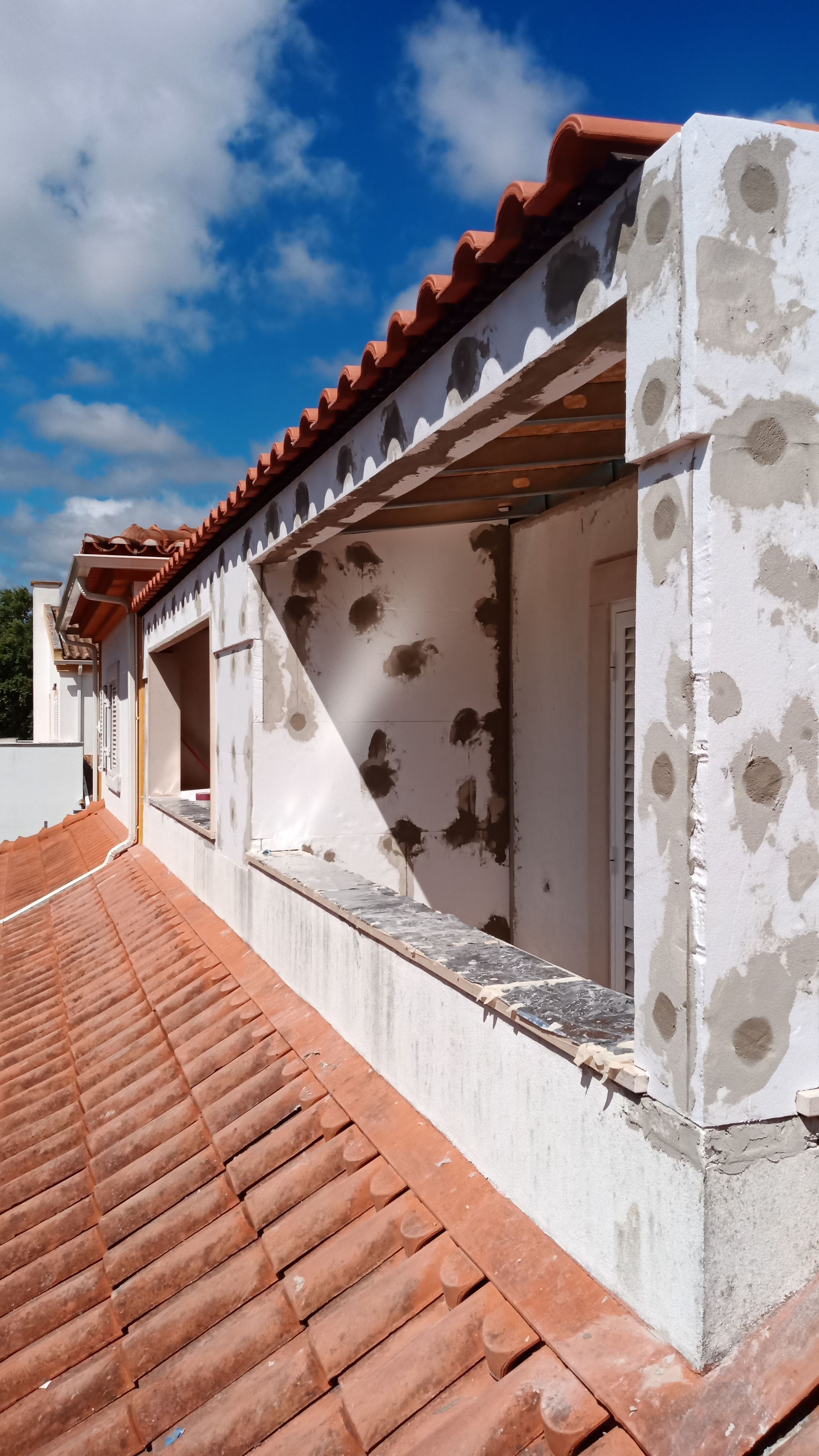 Exterior de um prédio em construção, com telhas, caixilhos de janelas e isolamento de isopor. Céu claro.