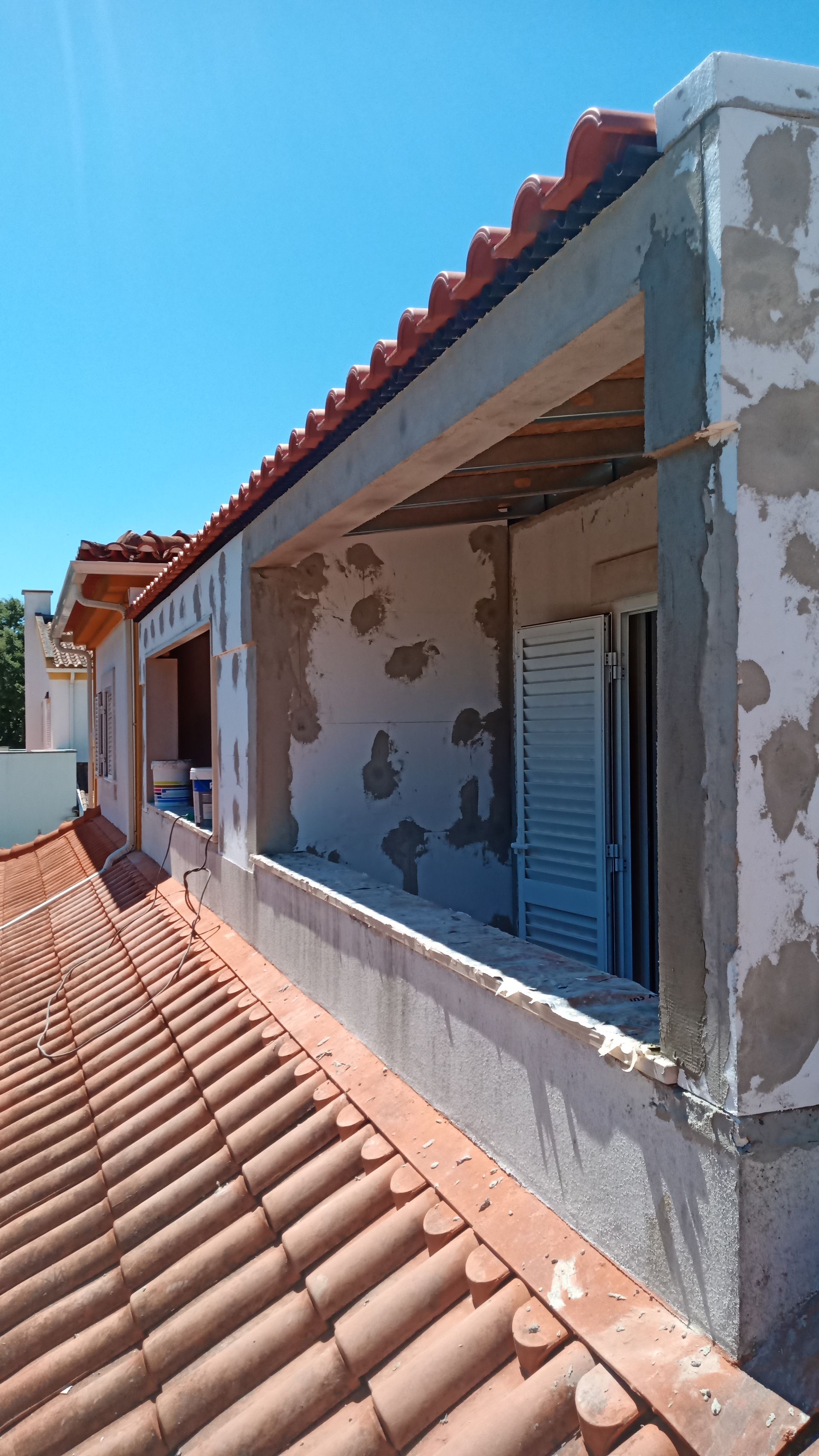 Vista do terraço de um prédio em construção. Telhado de telha vermelha, janelas abertas, paredes de gesso cinza, céu azul.
