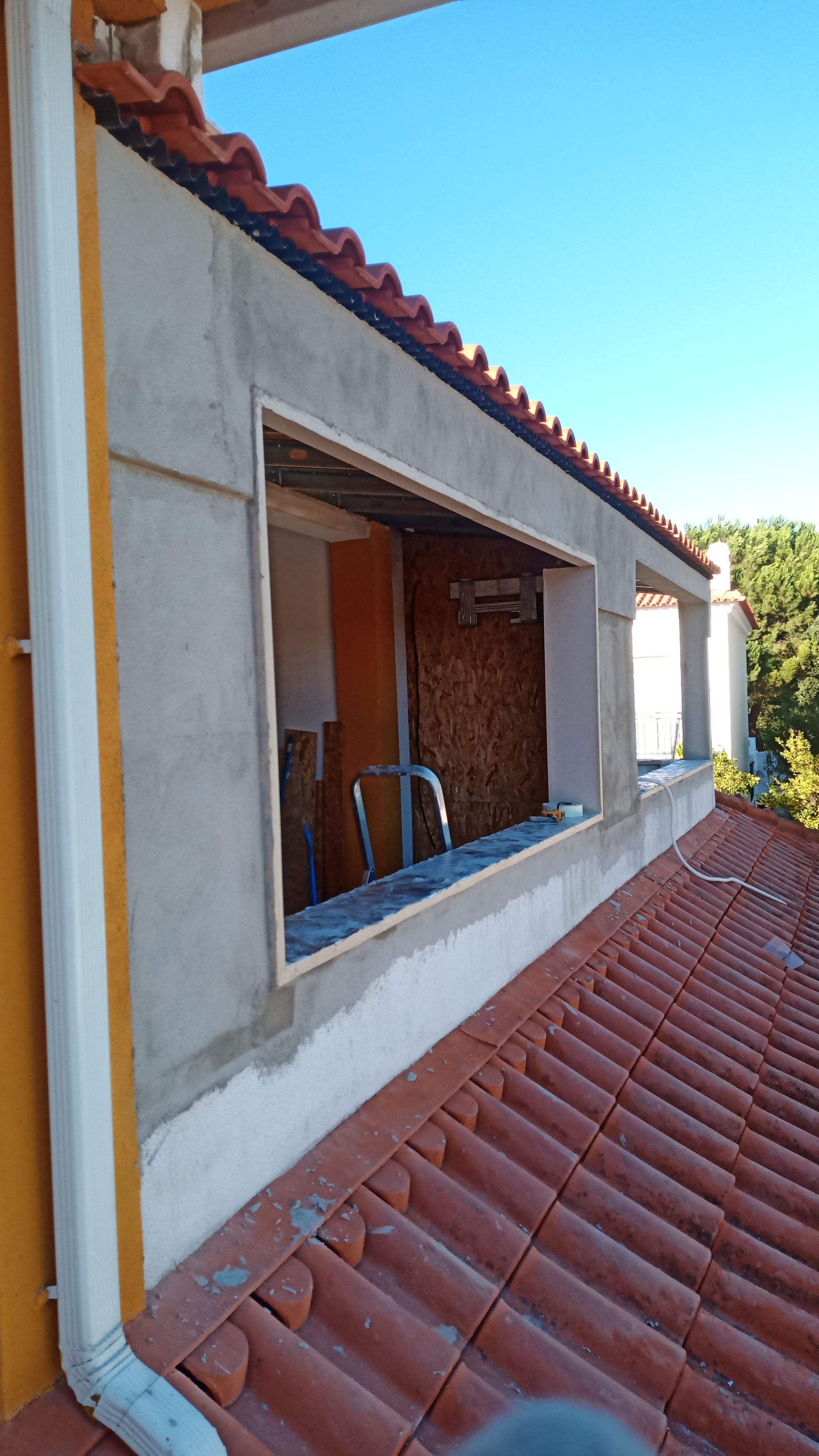 Vista exterior de um edifício em construção, mostrando a moldura de uma janela num telhado de telhas vermelhas e uma calha de drenagem.