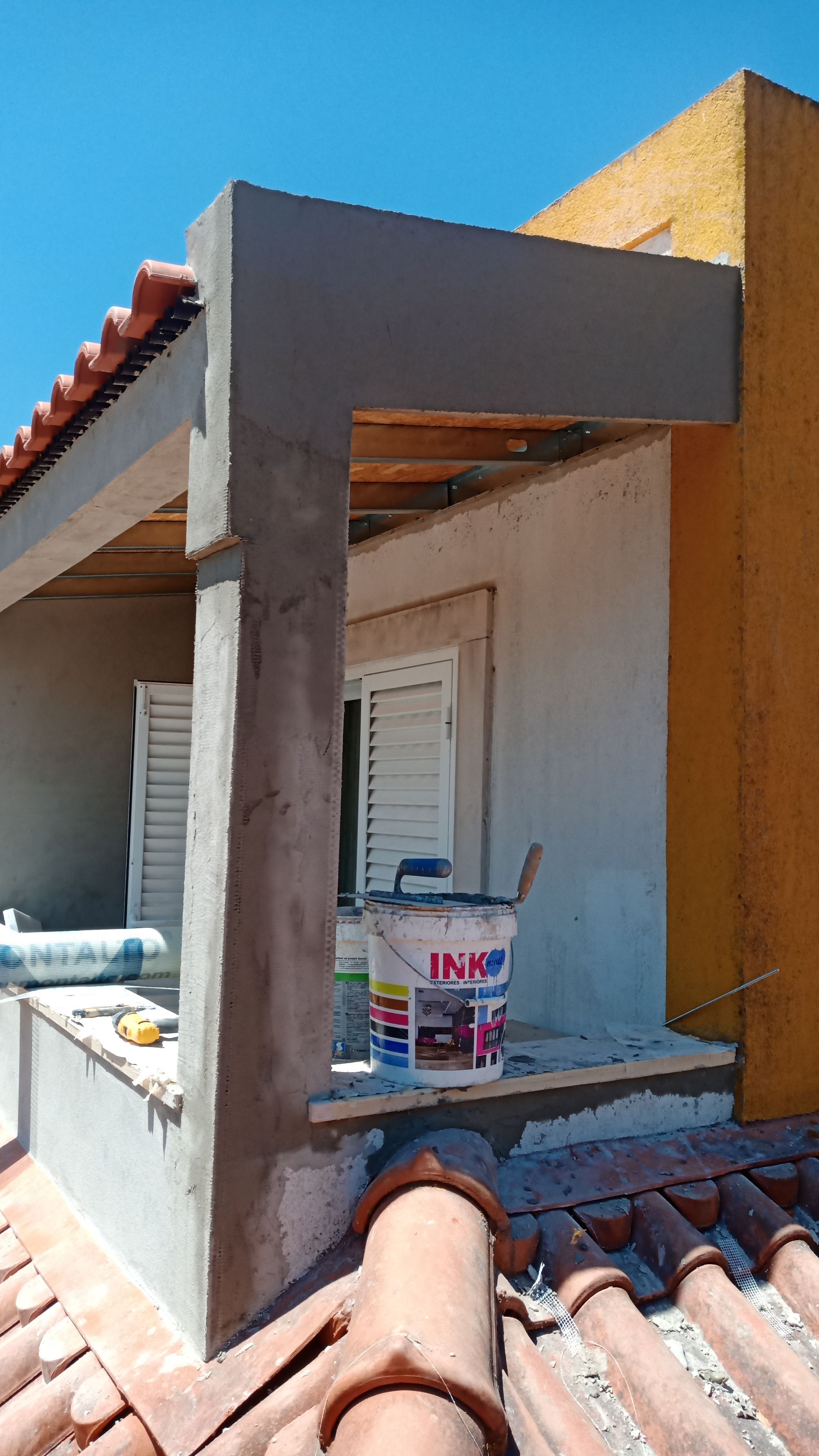 Vista exterior de um edifício em construção, mostrando telhas, uma viga de sustentação e um balde de tinta.