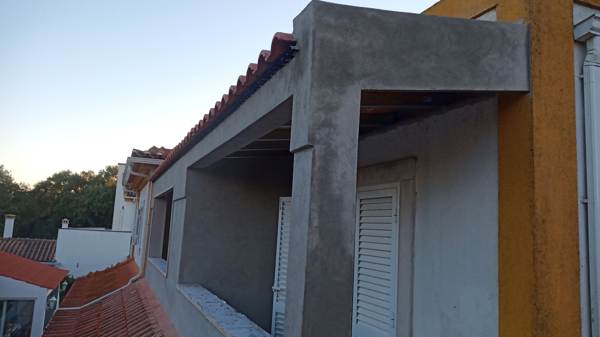 Exterior do edifício com beiral de concreto, telhado de telha vermelha e venezianas brancas.