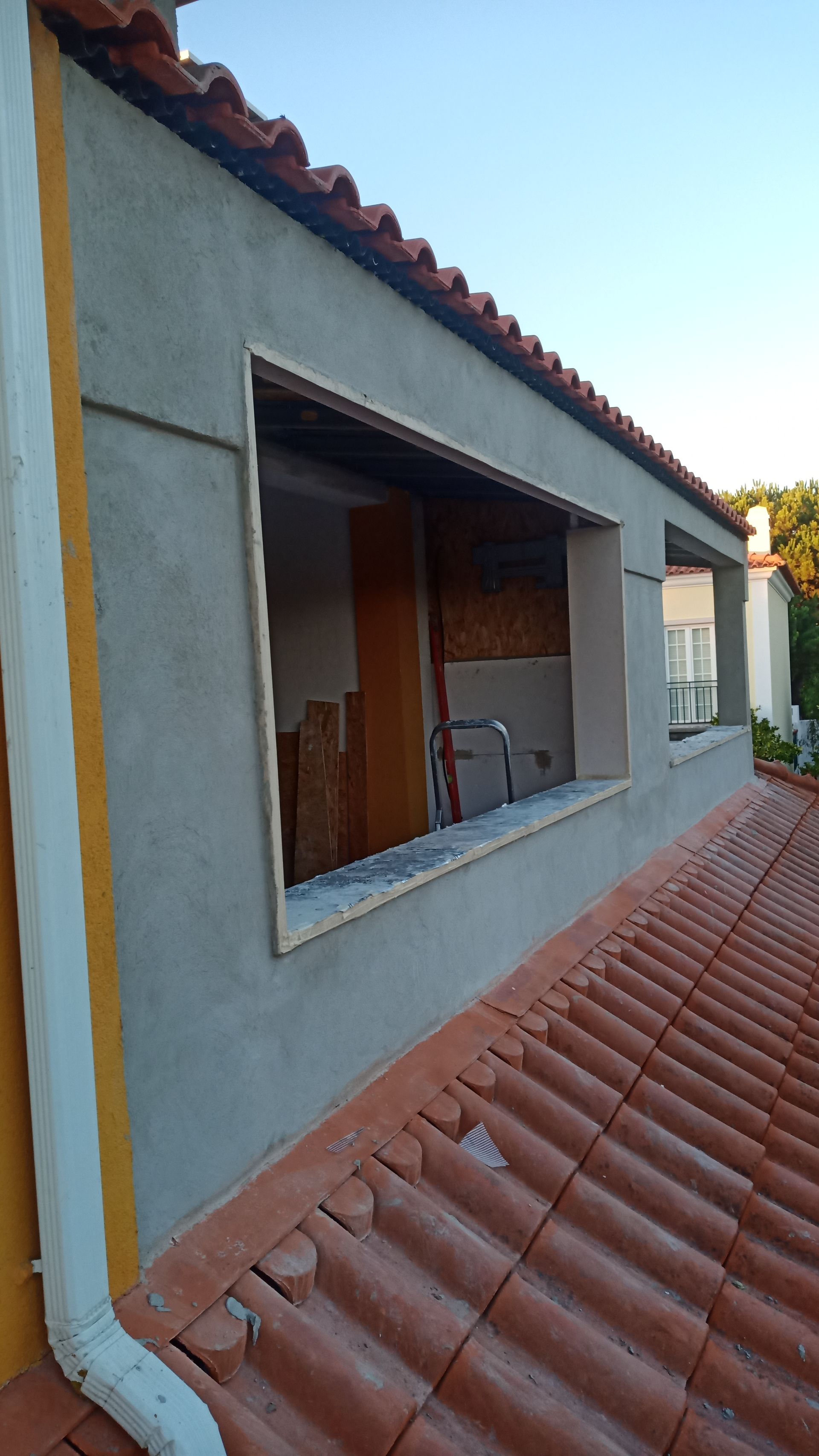 Parede externa com moldura de janela aberta, estuque cinza, telhado de telha vermelha e porta interna visível.