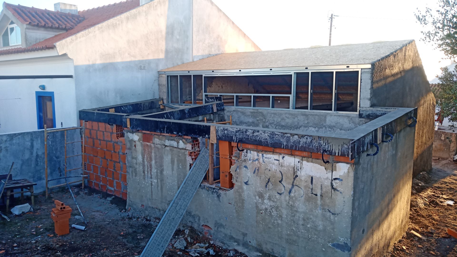 Canteiro de obras: construção com blocos de concreto, tijolos e estrutura metálica com telhado e caixilhos de janelas de metal.