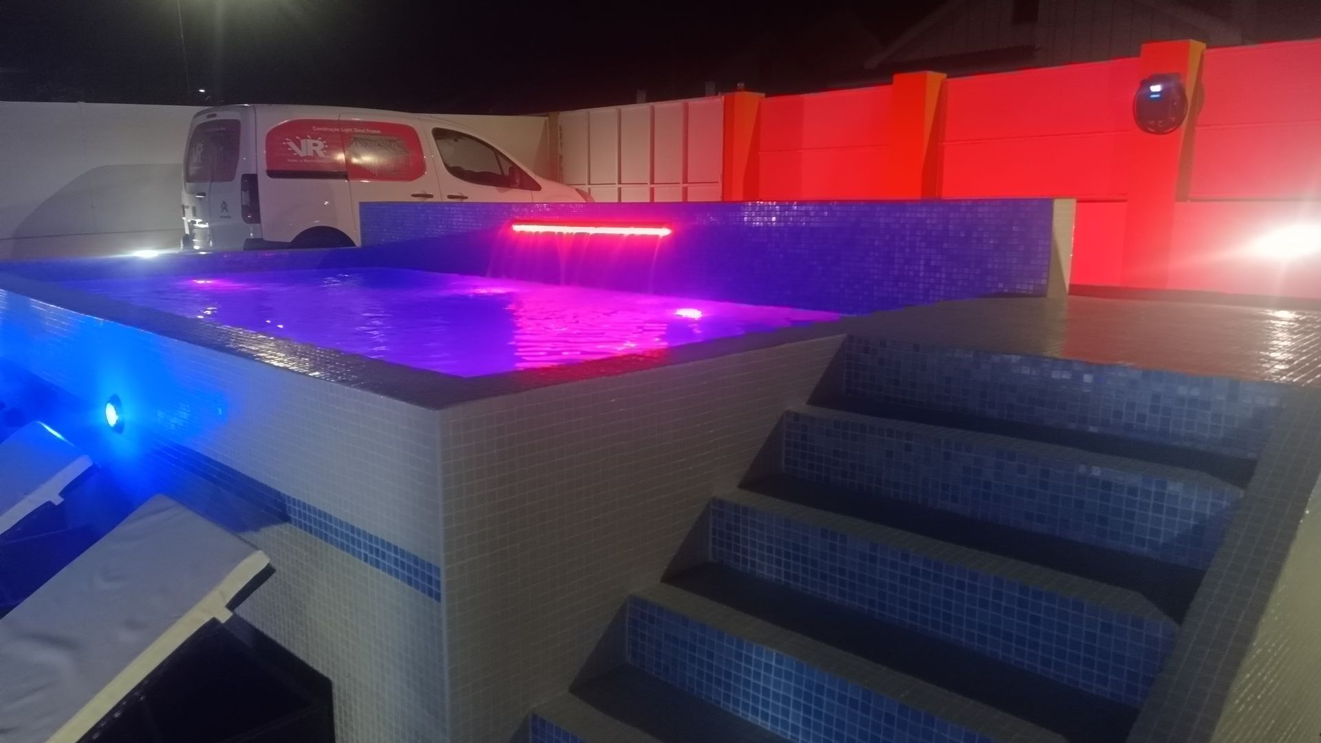 Piscina iluminada à noite com luzes azuis e rosas. Degraus levam à água. Uma van está estacionada nas proximidades.