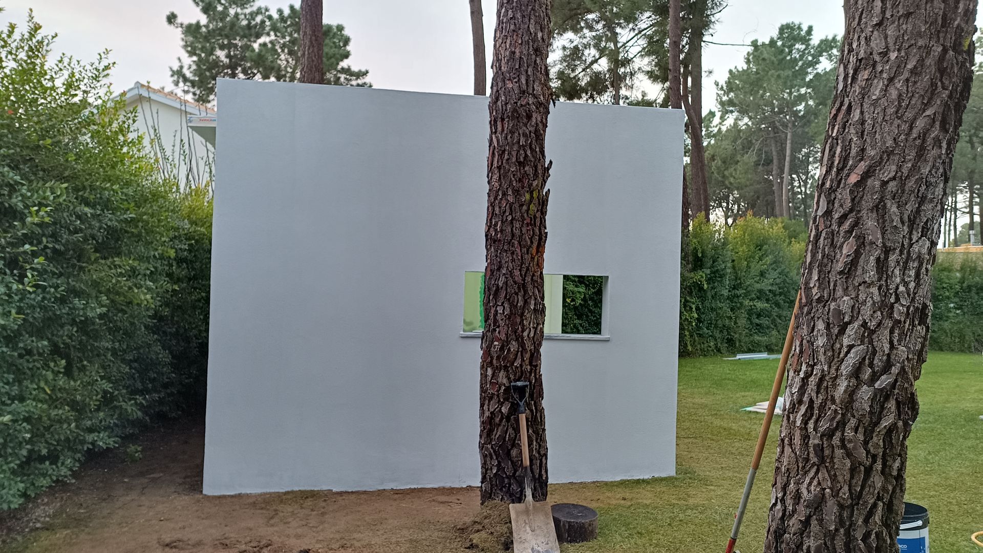 Parede branca com uma pequena janela entre duas árvores em um quintal gramado, cercado por vegetação.