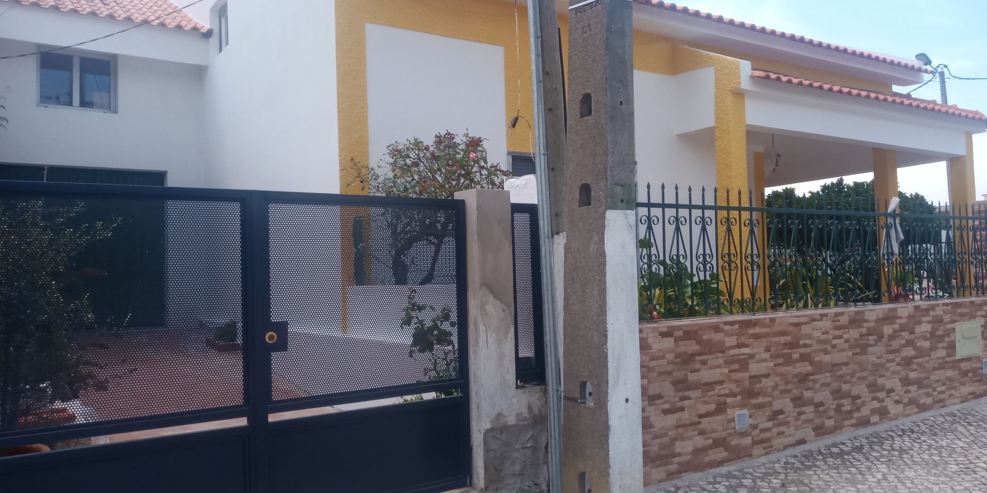 Uma casa com paredes brancas, detalhes em amarelo e telhado de telhas vermelhas. Um portão e uma cerca pretos ficam na frente.
