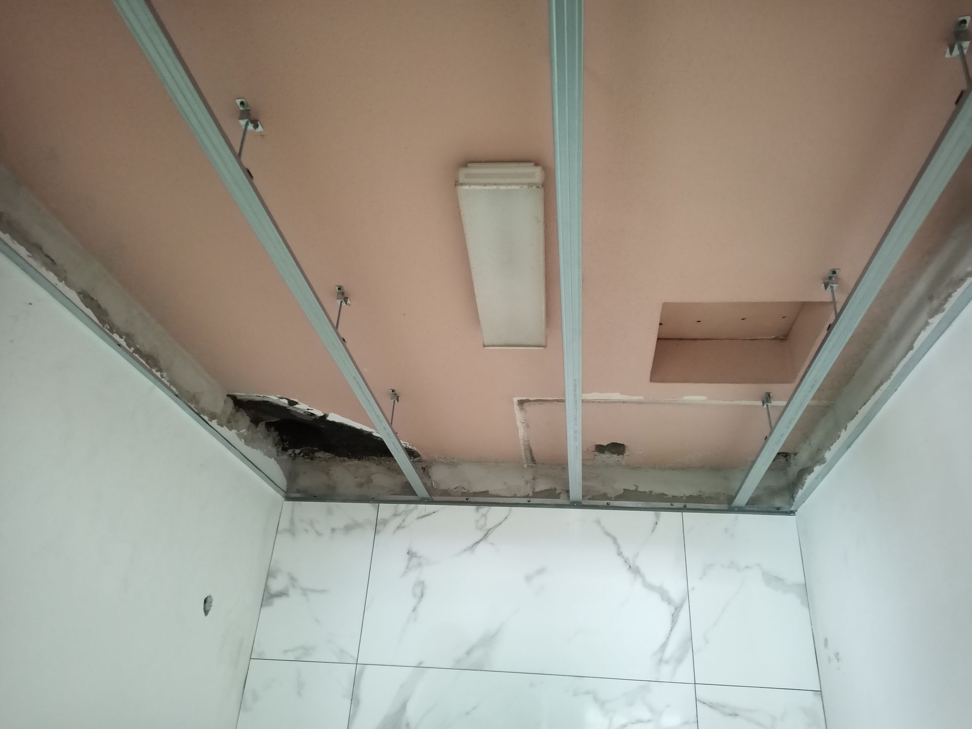 Ambiente interno com estrutura do teto aparente. Paredes de gesso rosa, suportes metálicos e área do teto danificada.