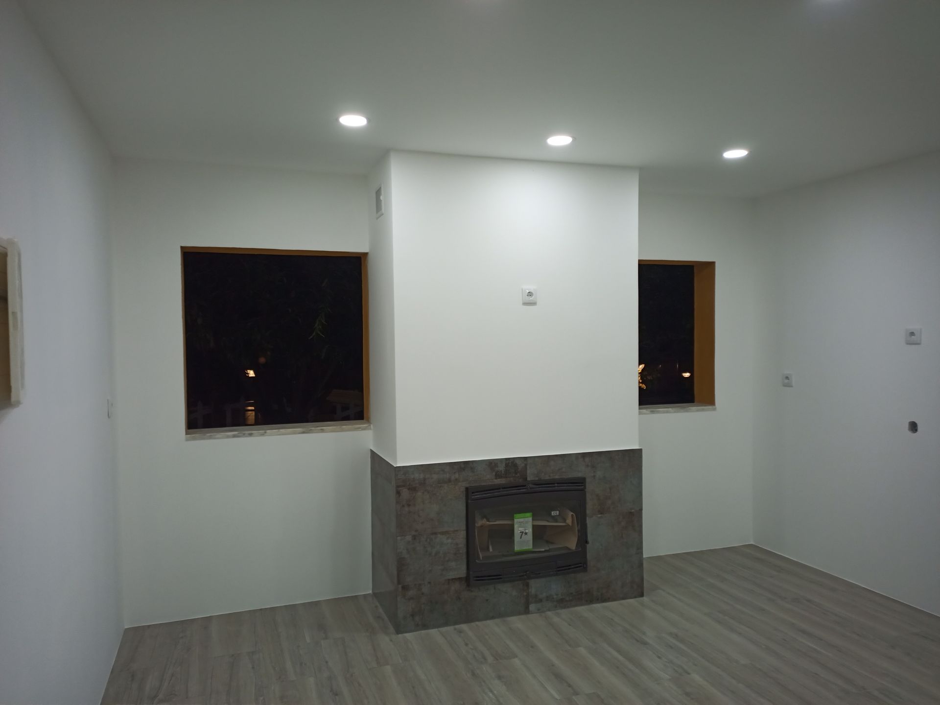 Sala interna com lareira branca, duas janelas e iluminação embutida. Piso cinza.