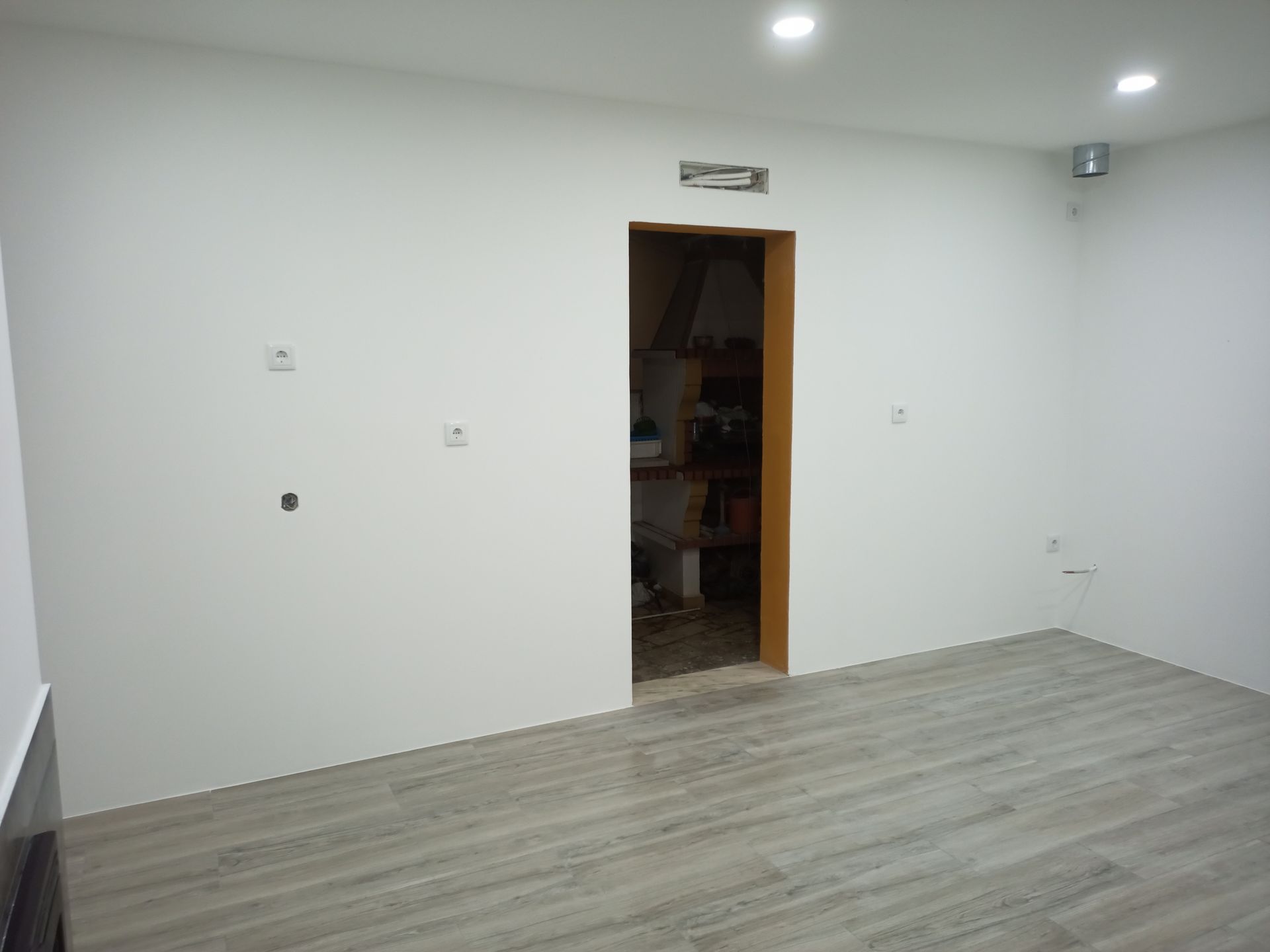 Quarto vazio com piso imitando madeira em tom cinza claro, paredes brancas e uma porta aberta que dá para um espaço desorganizado.