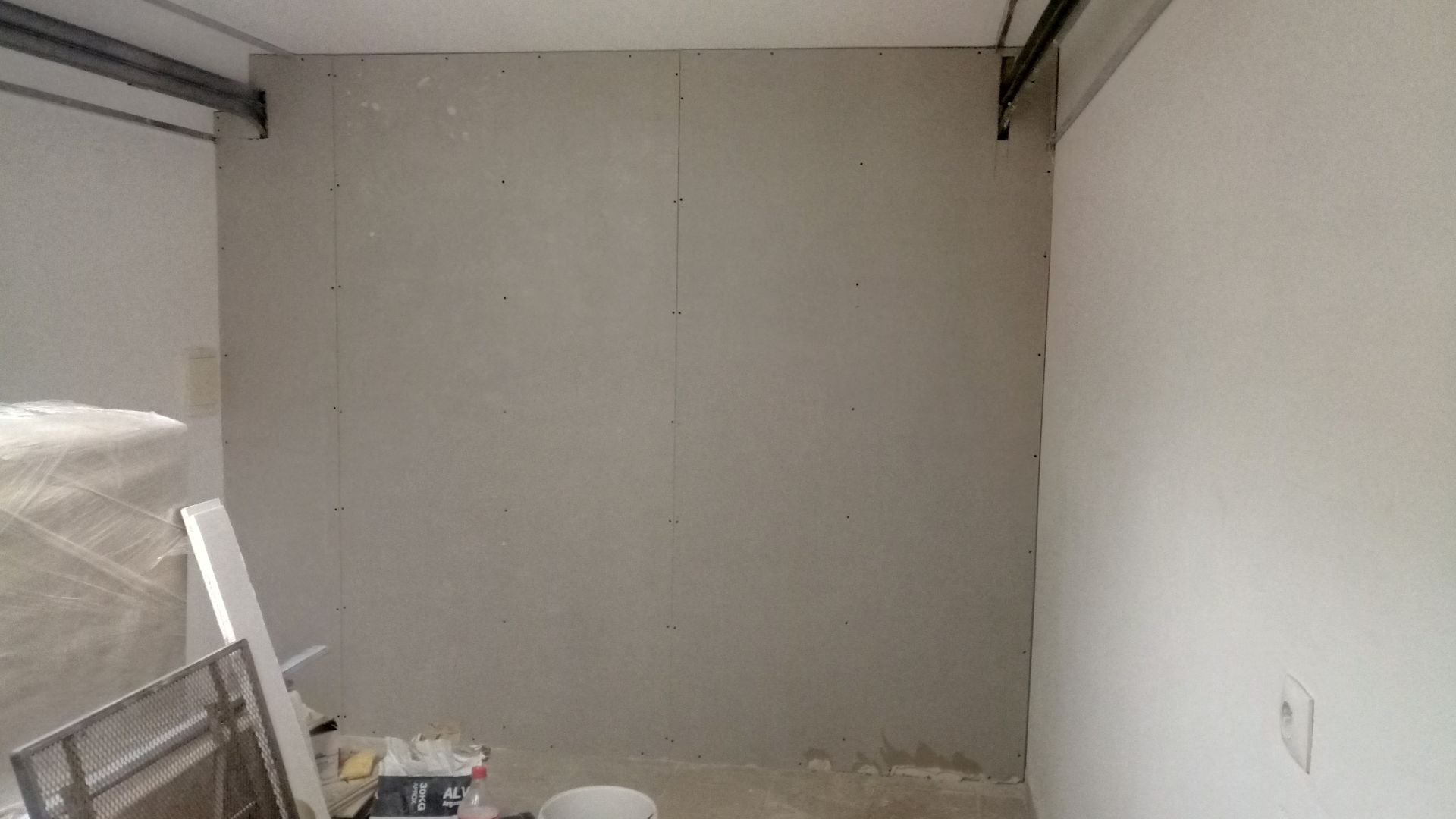 Sala vazia em construção, com paredes cinzentas, vigas metálicas e materiais de construção no chão.