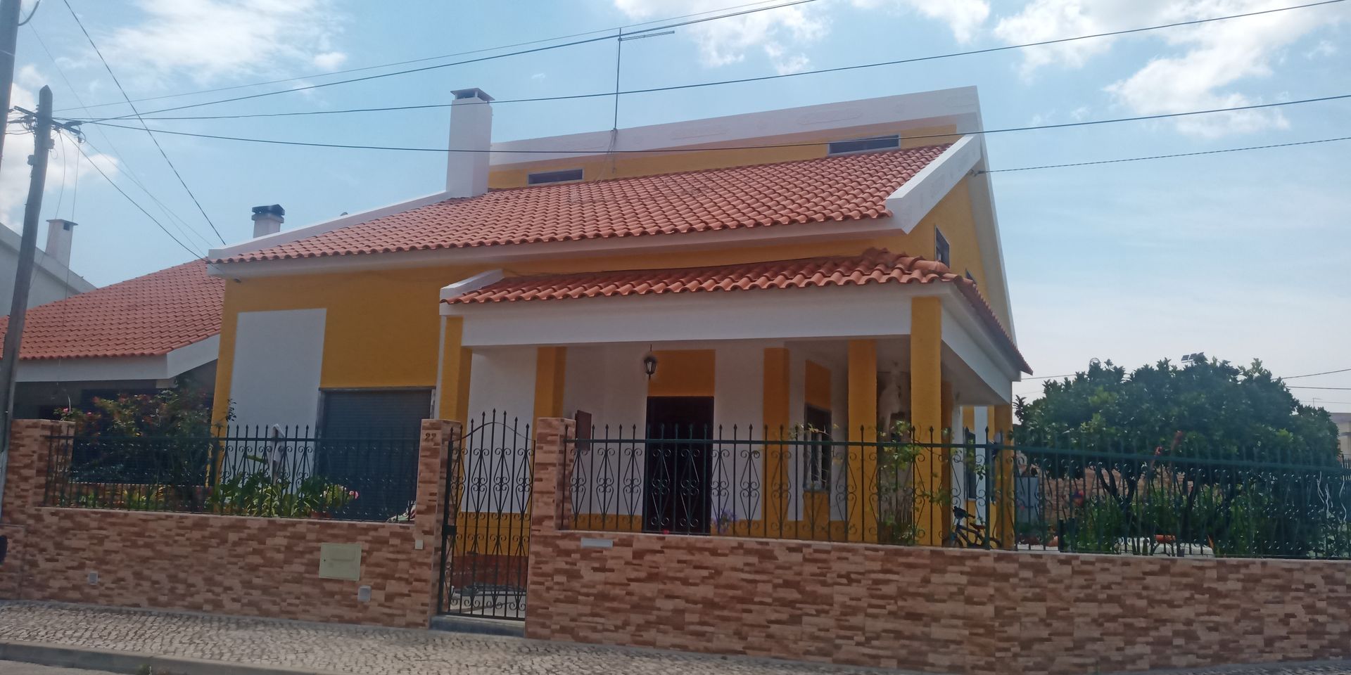 Casa amarela com telhado de telha vermelha, detalhes brancos e muro de tijolos. Cerca preta na frente.