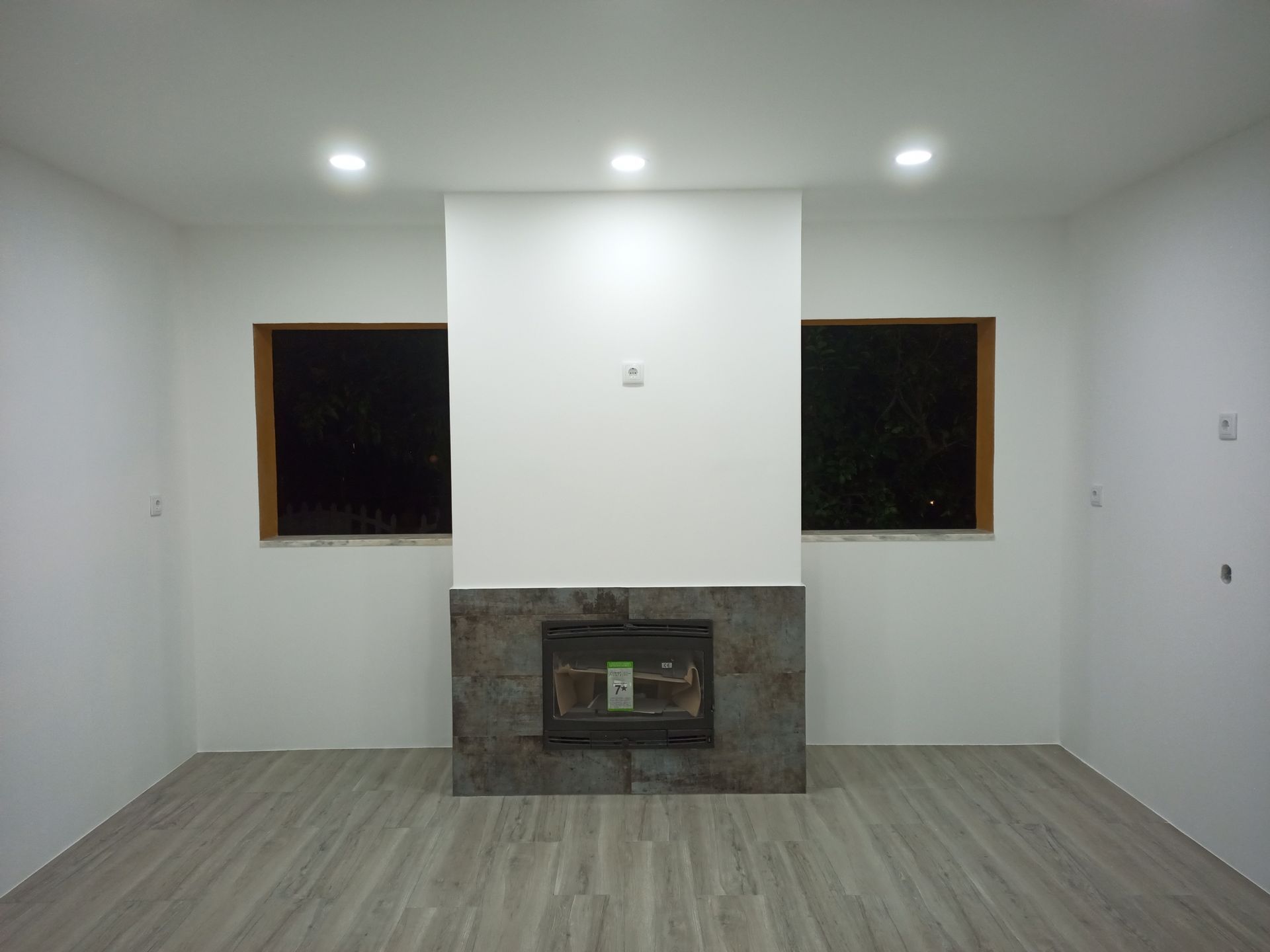 Sala vazia com lareira centralizada em uma parede. Duas janelas ladeiam a lareira, e o piso tem um acabamento que imita madeira.
