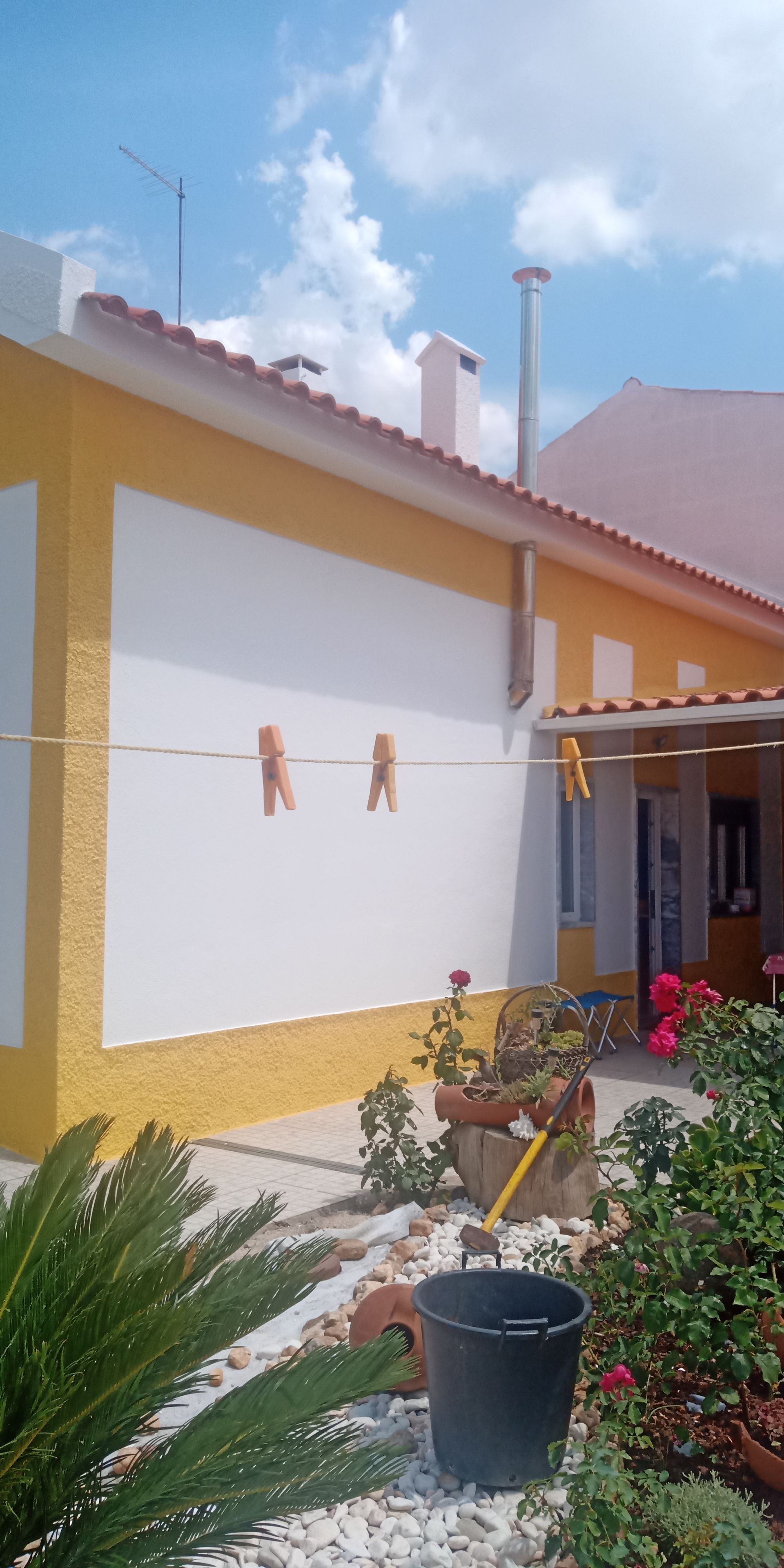 Casa amarela e branca com varal. Jardim com flores e um balde. Céu azul.