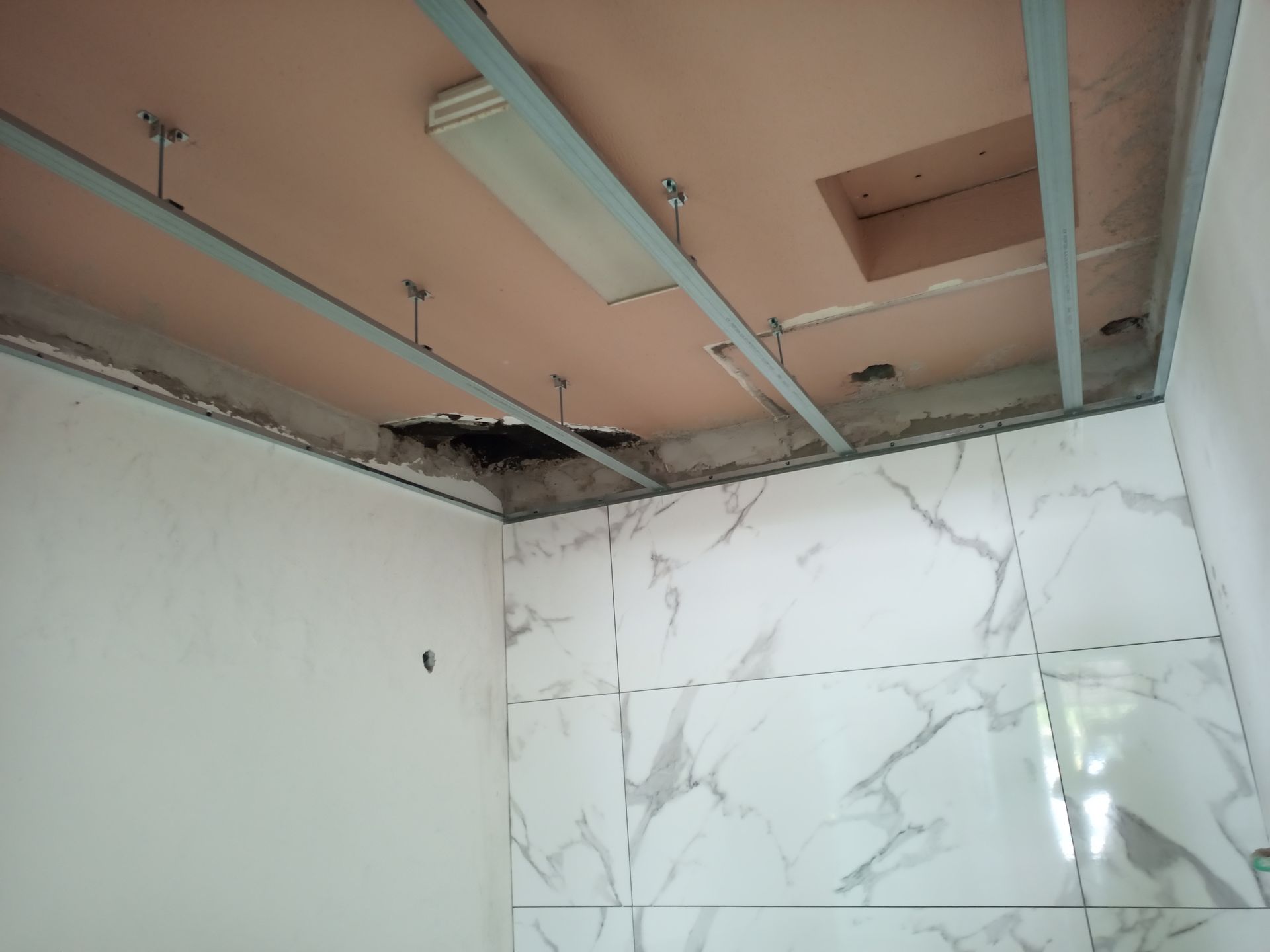 Reforma de banheiro com teto incompleto, estrutura metálica exposta e azulejos com aparência de mármore nas paredes.