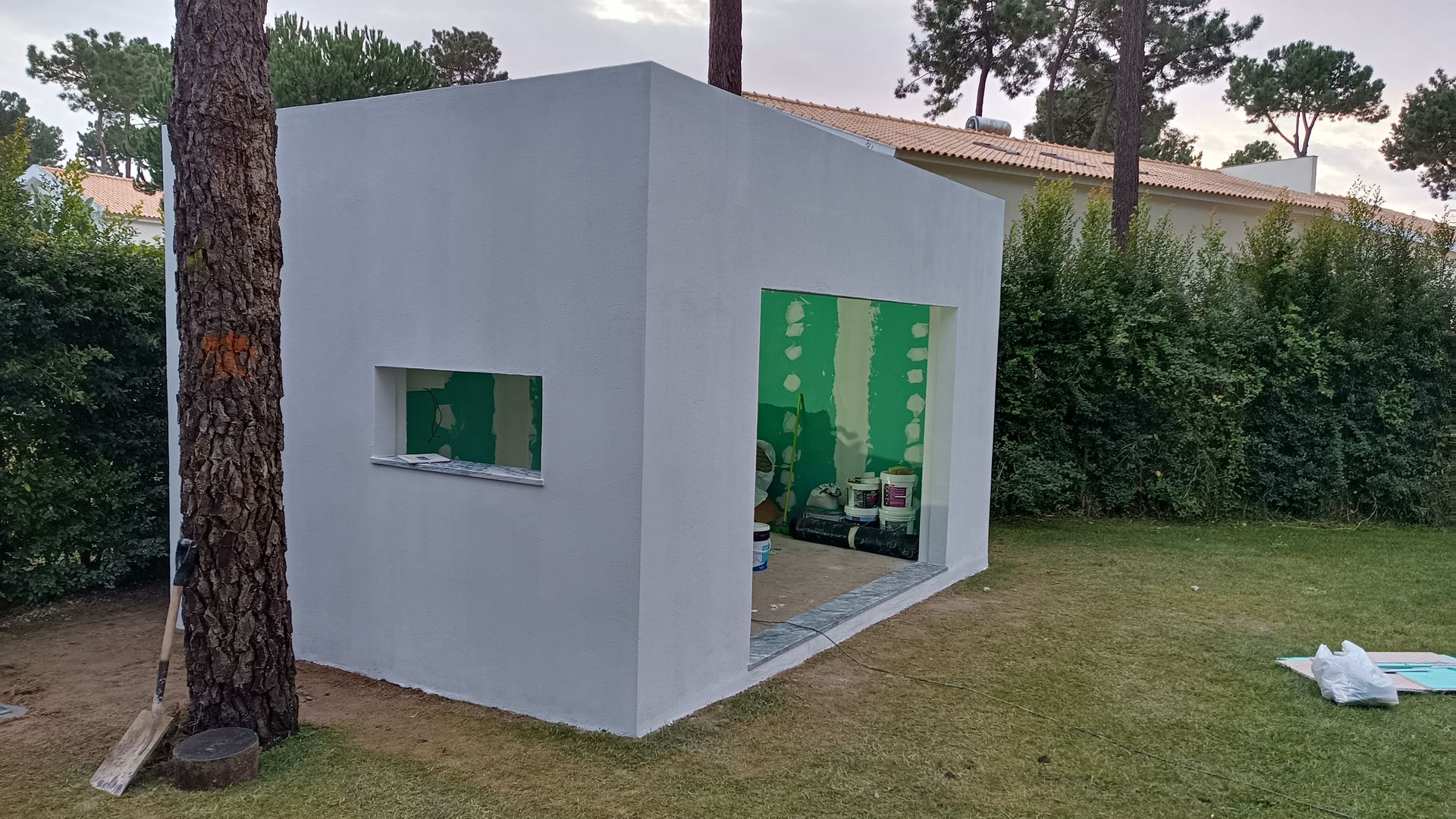 Estrutura externa branca e inacabada com janela e porta, cercada por grama, árvores e arbustos.