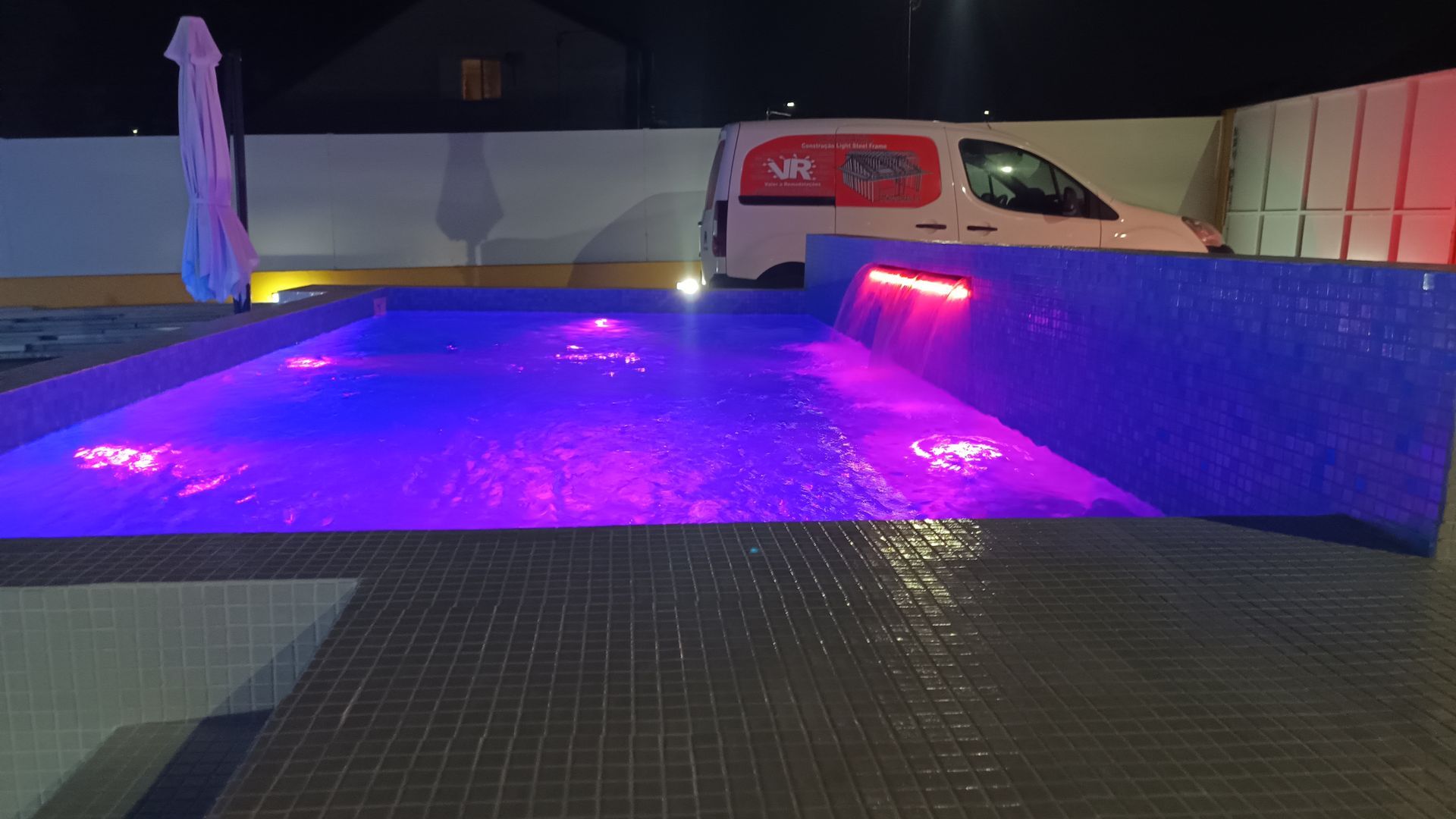 Vista noturna de uma piscina com iluminação roxa e uma cascata, com uma van branca ao fundo.
