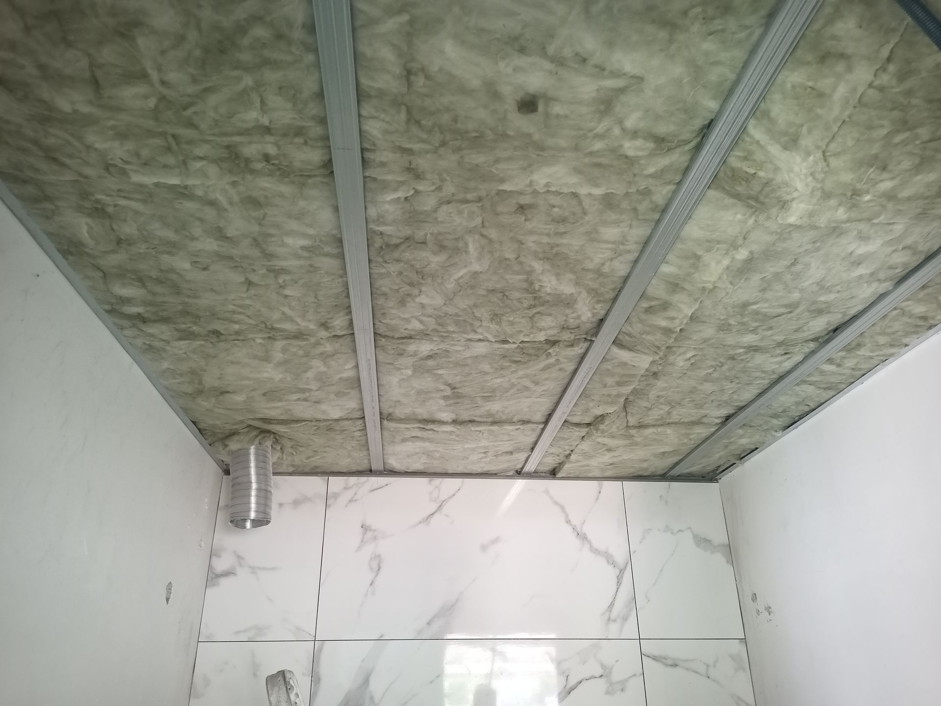 Teto com isolamento entre suportes metálicos em uma sala com paredes revestidas de azulejos.