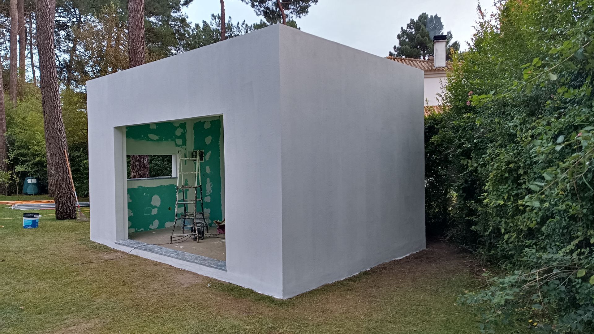 Prédio quadrado branco com porta e janela abertas, parede de gesso verde visível no interior, sobre grama.