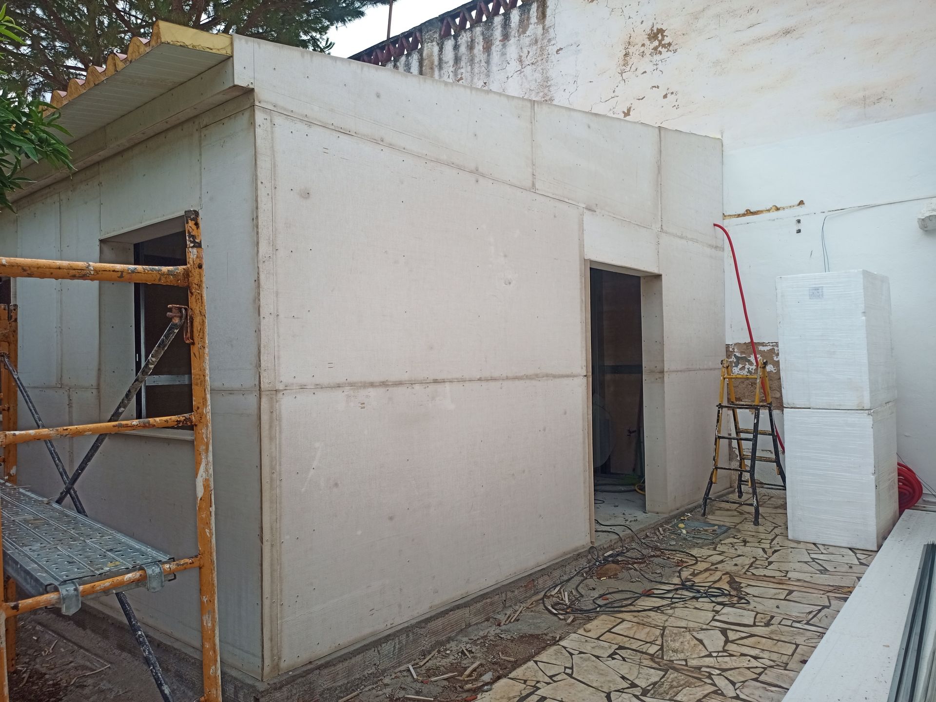 Prédio em construção, painéis de cor clara, andaimes, porta, janela, pequena estrutura externa.
