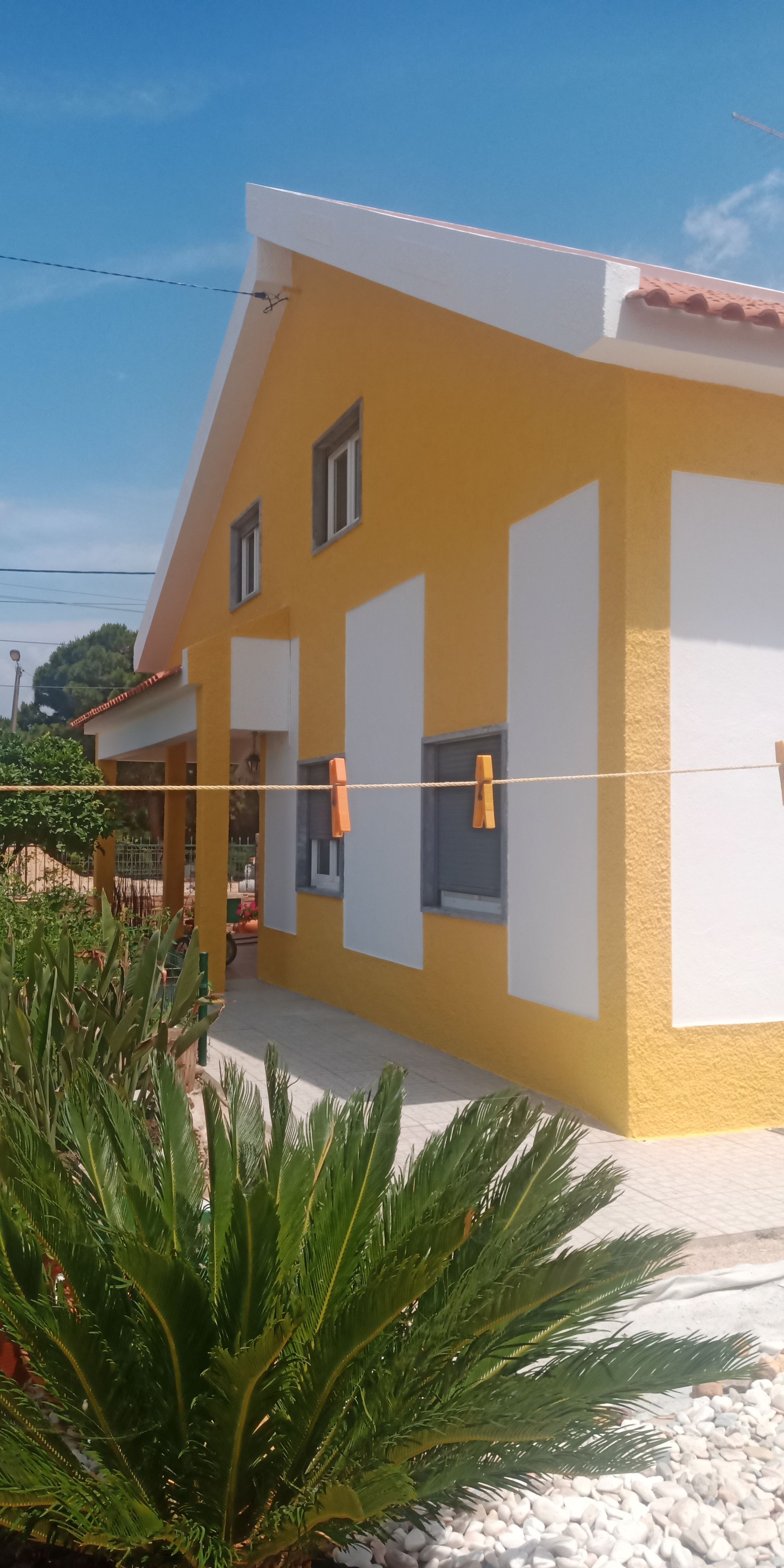 Casa amarela com detalhes em branco, céu azul e plantas verdes.