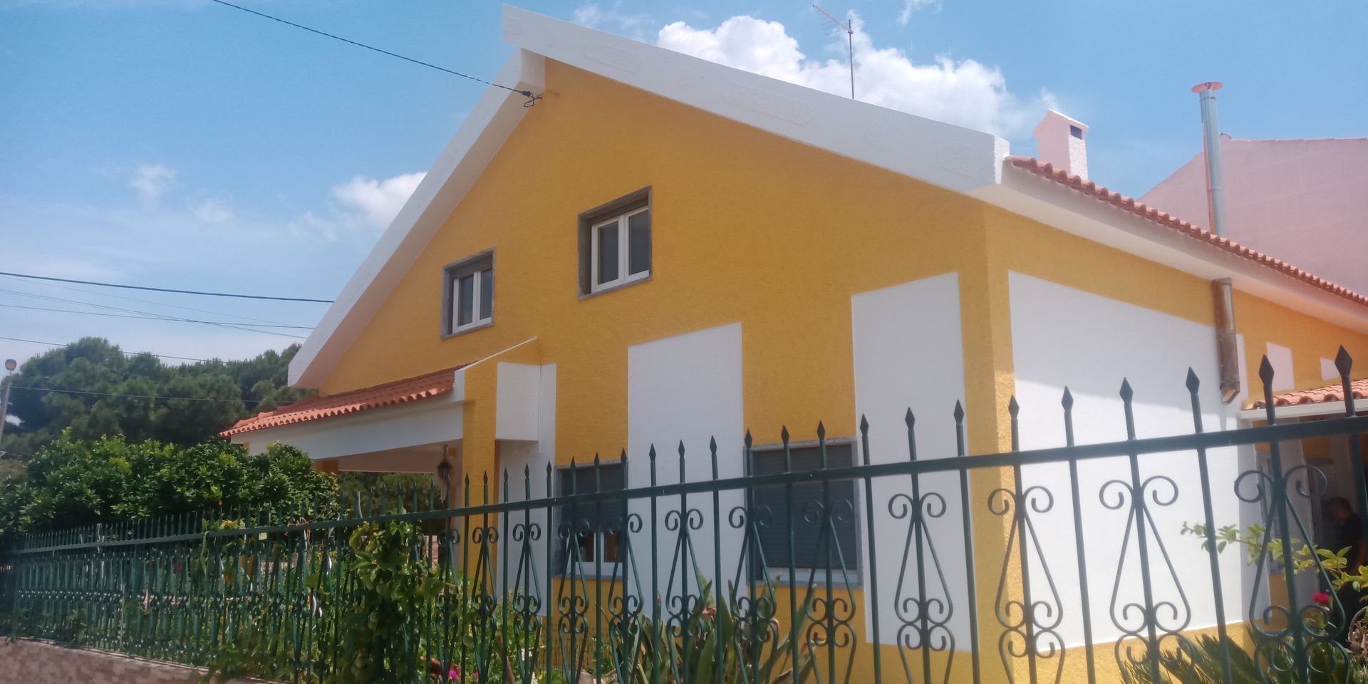 Casa amarela com detalhes brancos, cerca de ferro preta e céu azul.