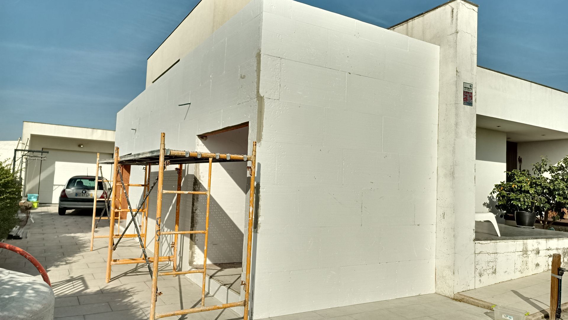 Vista exterior de um edifício em construção, com painéis de espuma branca numa parede, andaimes e um carro na entrada.