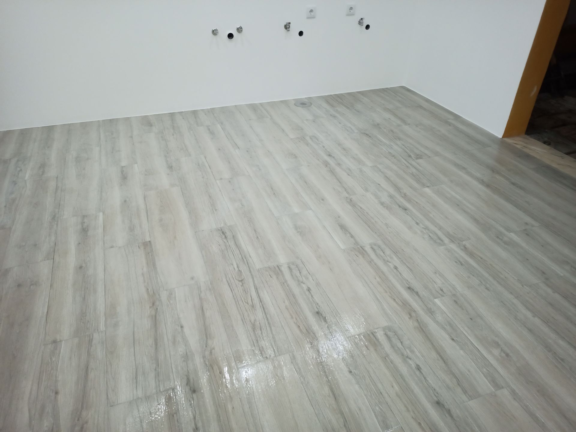 Piso com aparência de madeira clara em um cômodo com paredes brancas, tomadas elétricas inacabadas e uma porta.