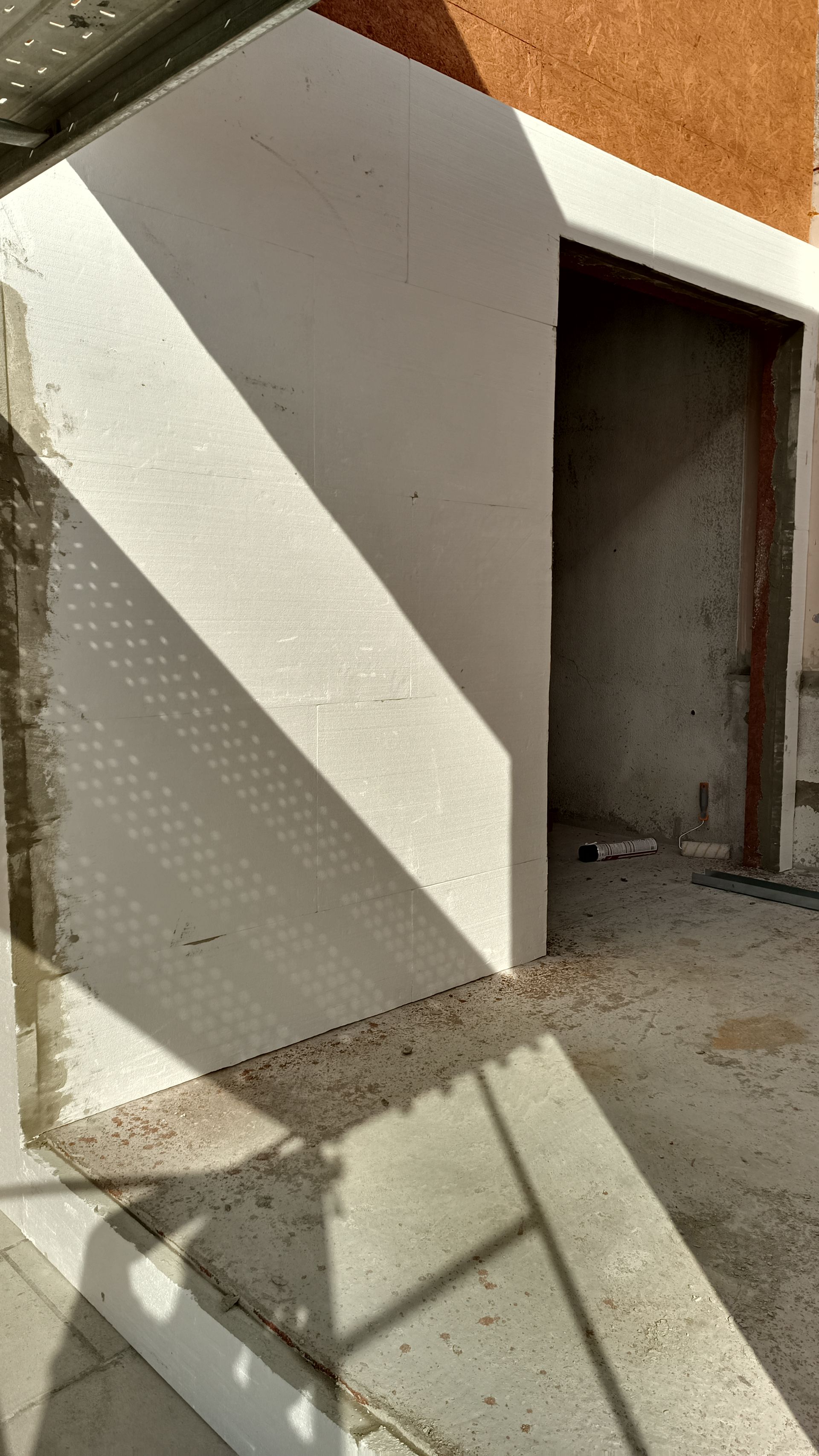 Canteiro de obras com parede e porta inacabadas, mostrando os materiais expostos e os padrões de luz e sombra.