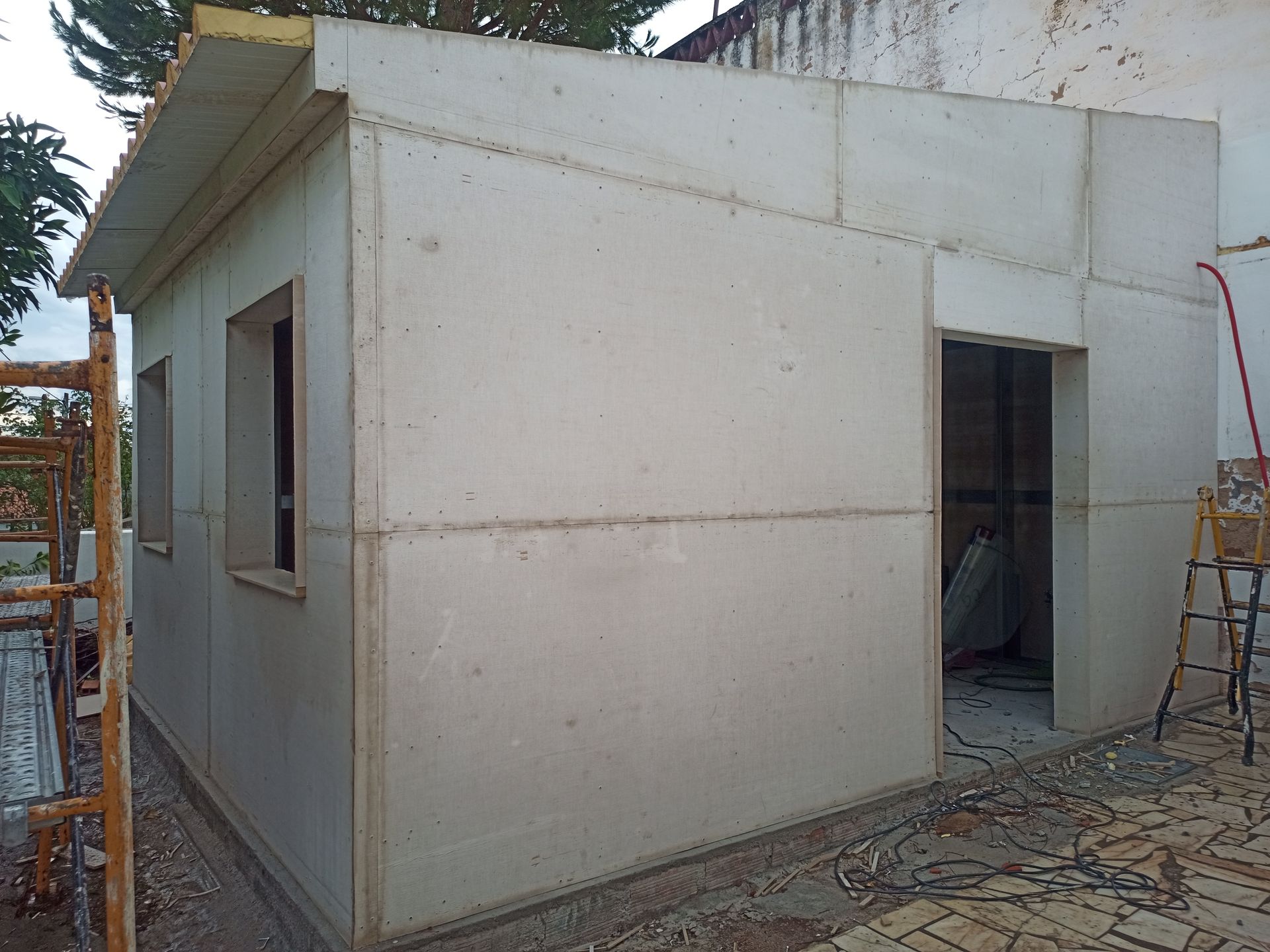 Construção de um pequeno edifício de concreto branco com aberturas para janelas e portas; ambiente externo.