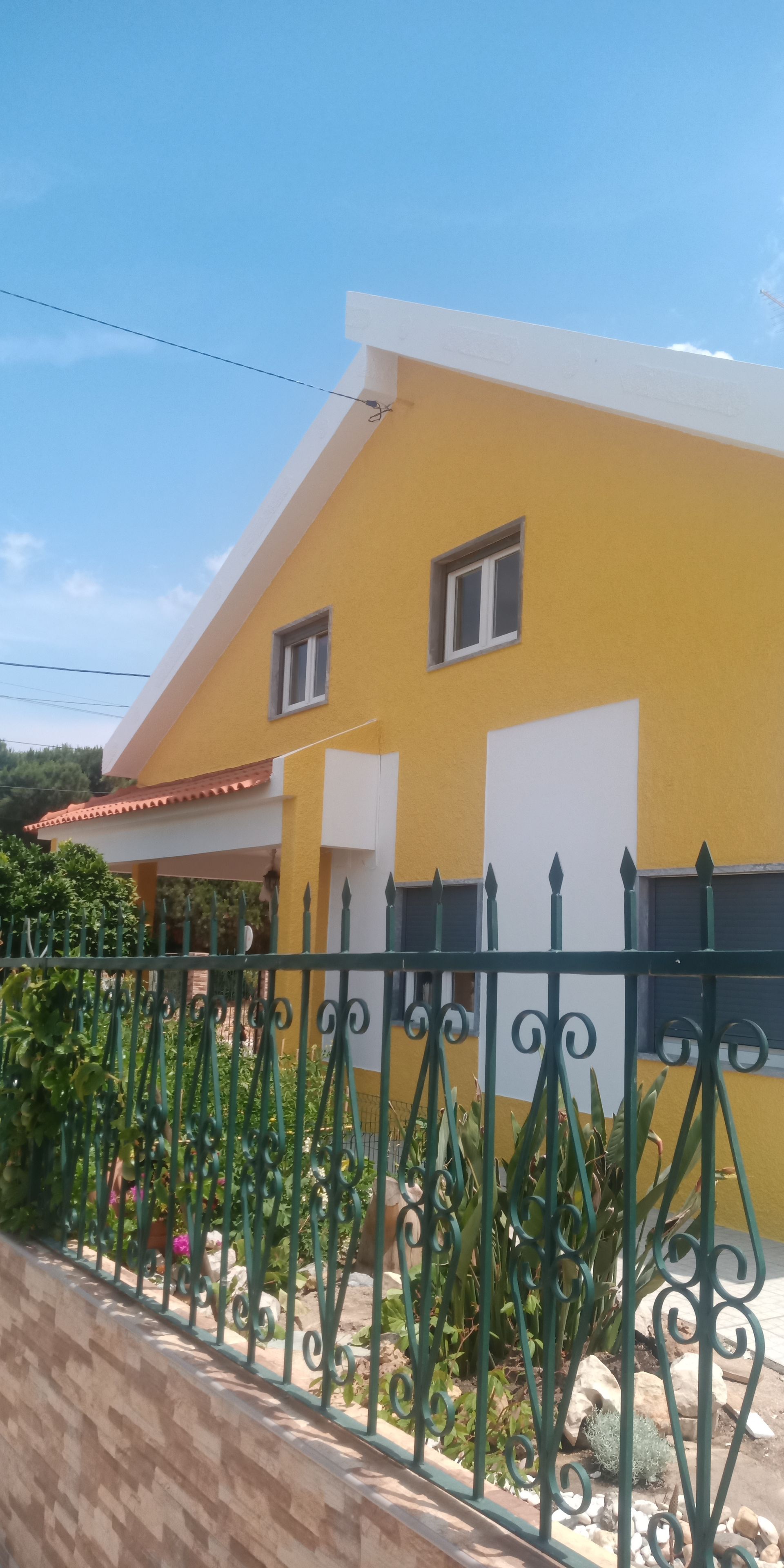 Casa amarela com detalhes, janelas e telhado brancos. Cerca verde na frente. Céu azul e limpo.