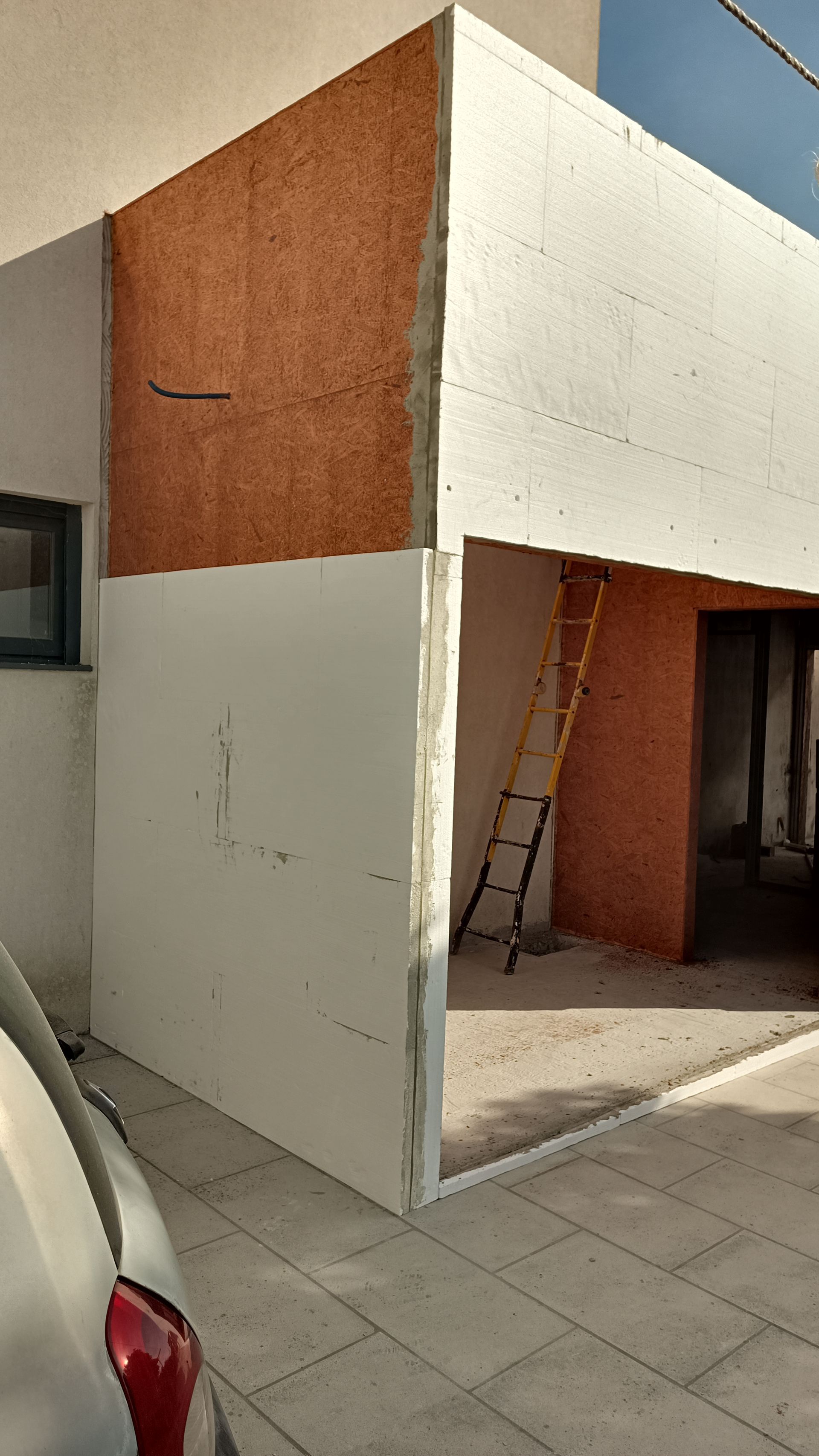 Exterior do prédio em construção, isolamento de espuma branca, painéis de madeira marrom, escada amarela no interior.