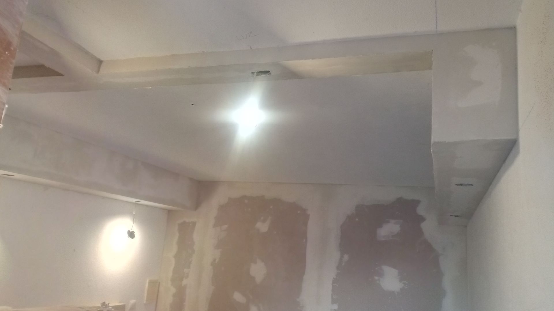 Quarto em construção; drywall no teto e nas paredes; vigas expostas; luz forte no centro.