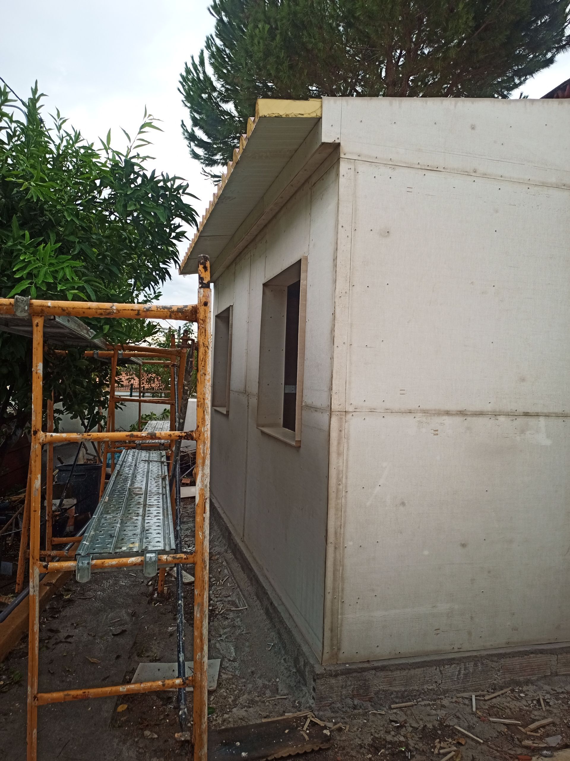 Exterior de um edifício de concreto em construção com duas janelas, uma árvore e andaimes.
