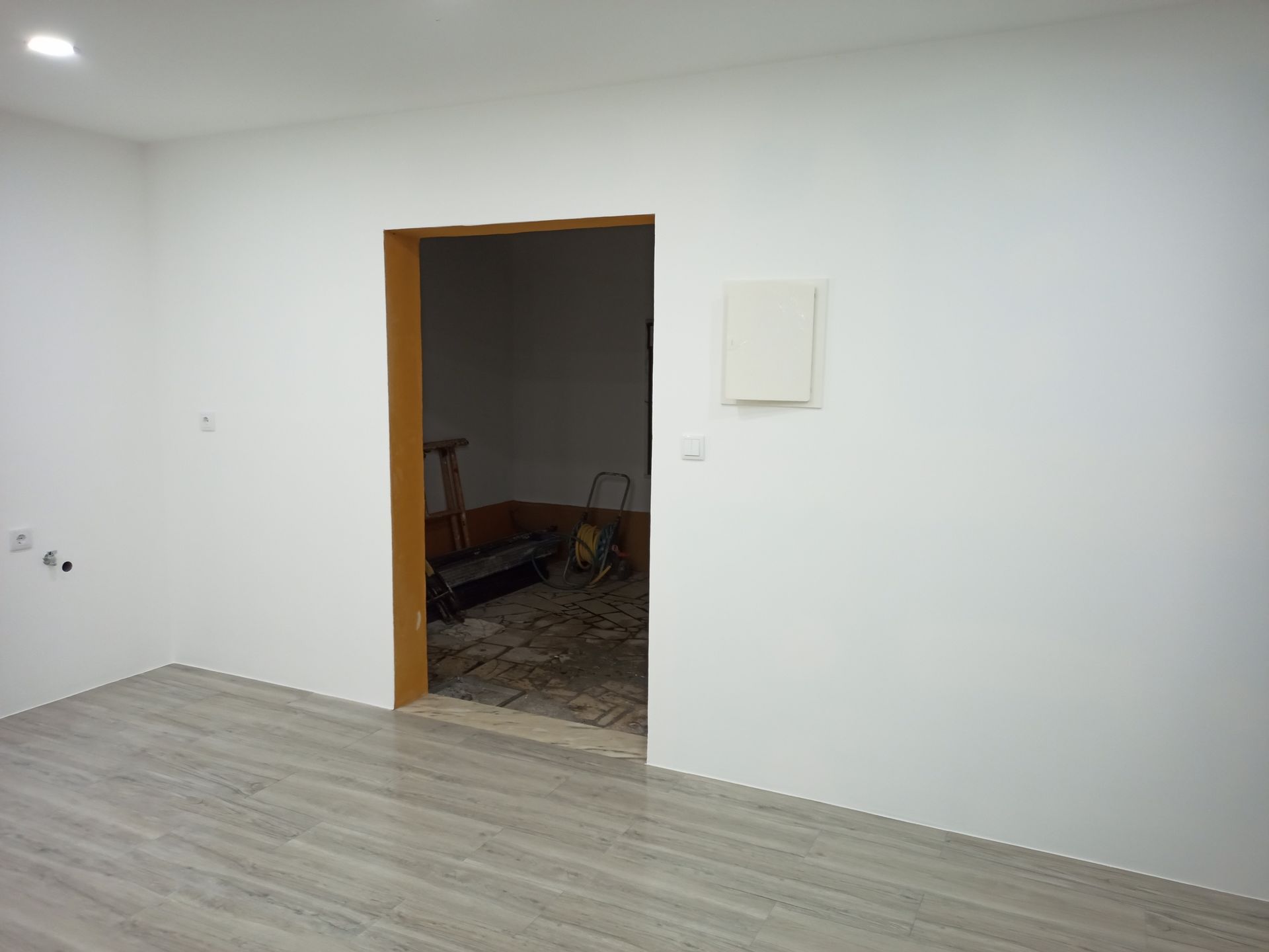 Quarto com paredes brancas e porta que dá para outro cômodo. Piso de madeira clara e piso inacabado.