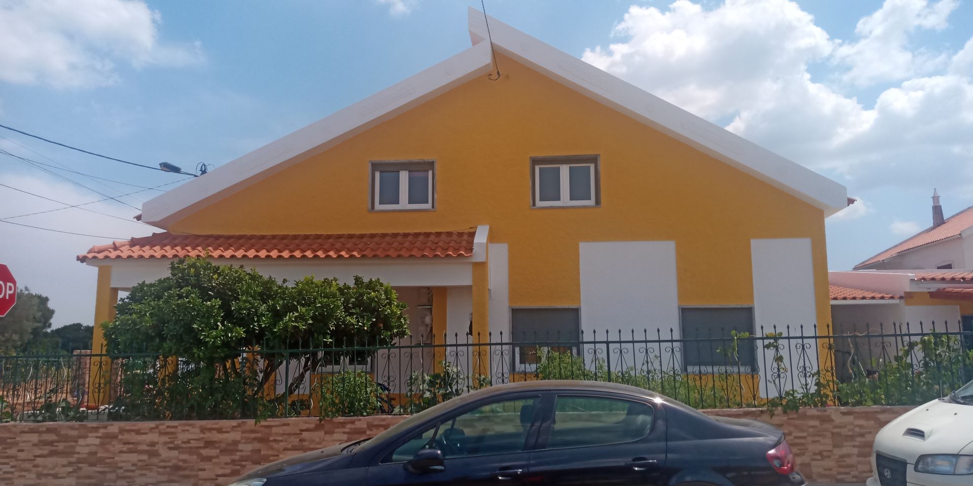 Casa amarela com detalhes brancos e telhado vermelho. Um carro está estacionado em frente à casa. Céu azul.