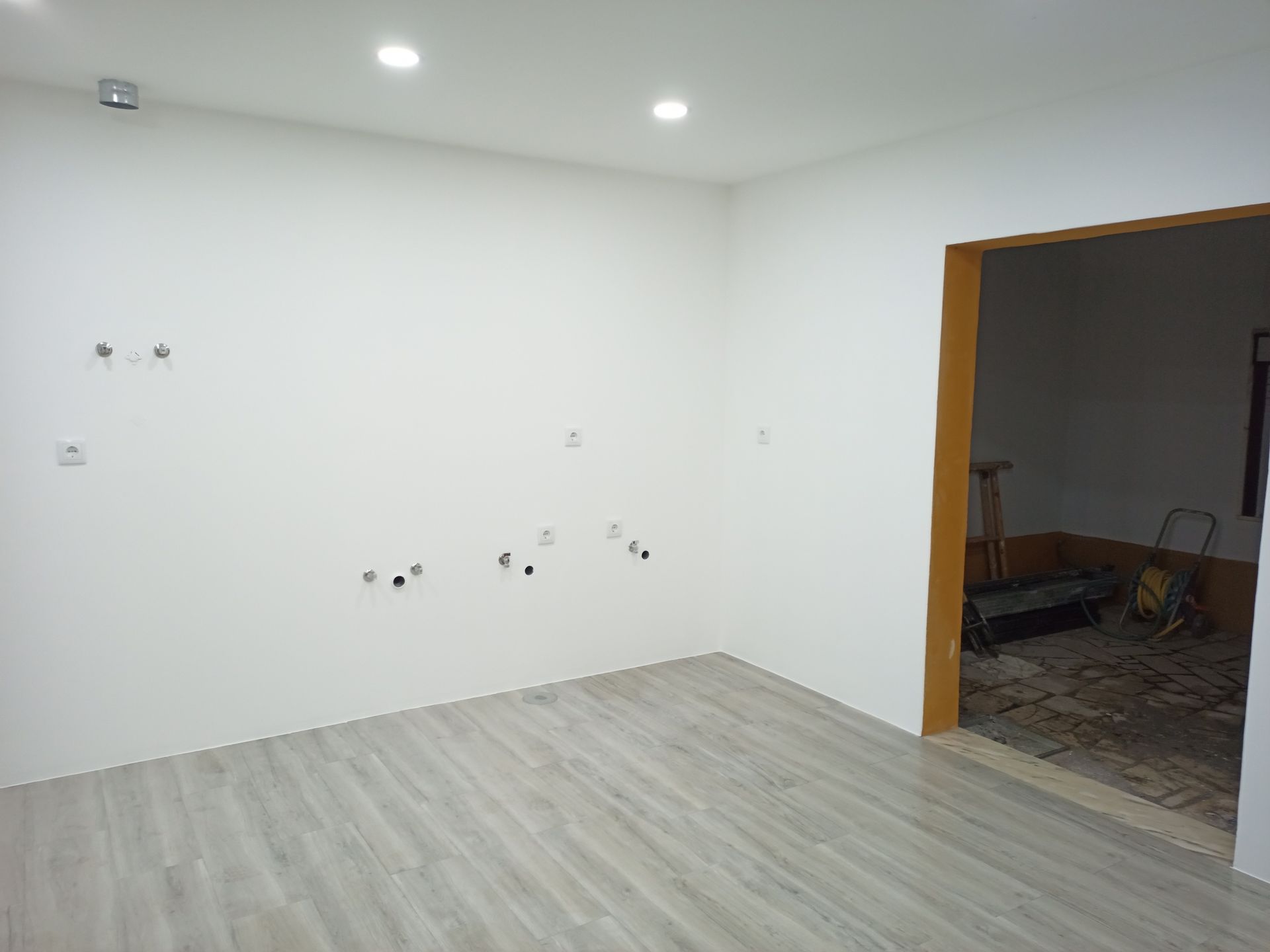 Quarto com paredes brancas, piso de madeira em tons de cinza, porta aberta e tomadas e acessórios elétricos.