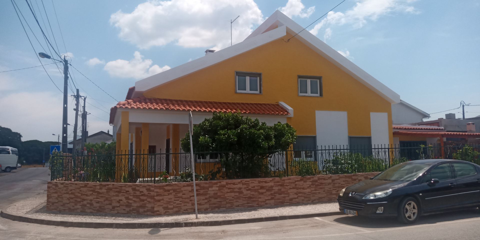 Casa amarela com telhado branco, parede de tijolos marrons e um carro preto estacionado em frente.