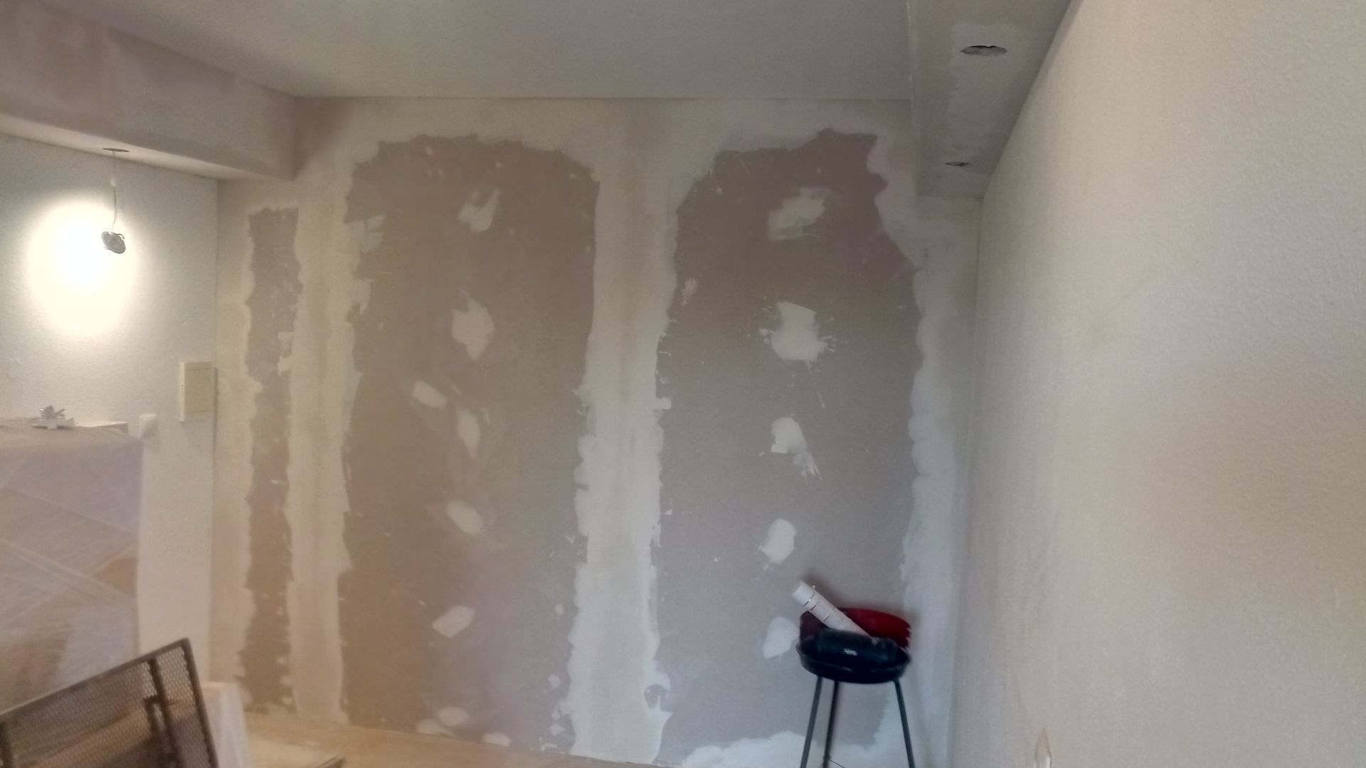 Construção de parede interna com drywall, parcialmente acabada. Apresenta manchas de massa corrida, ferramentas e um banquinho.
