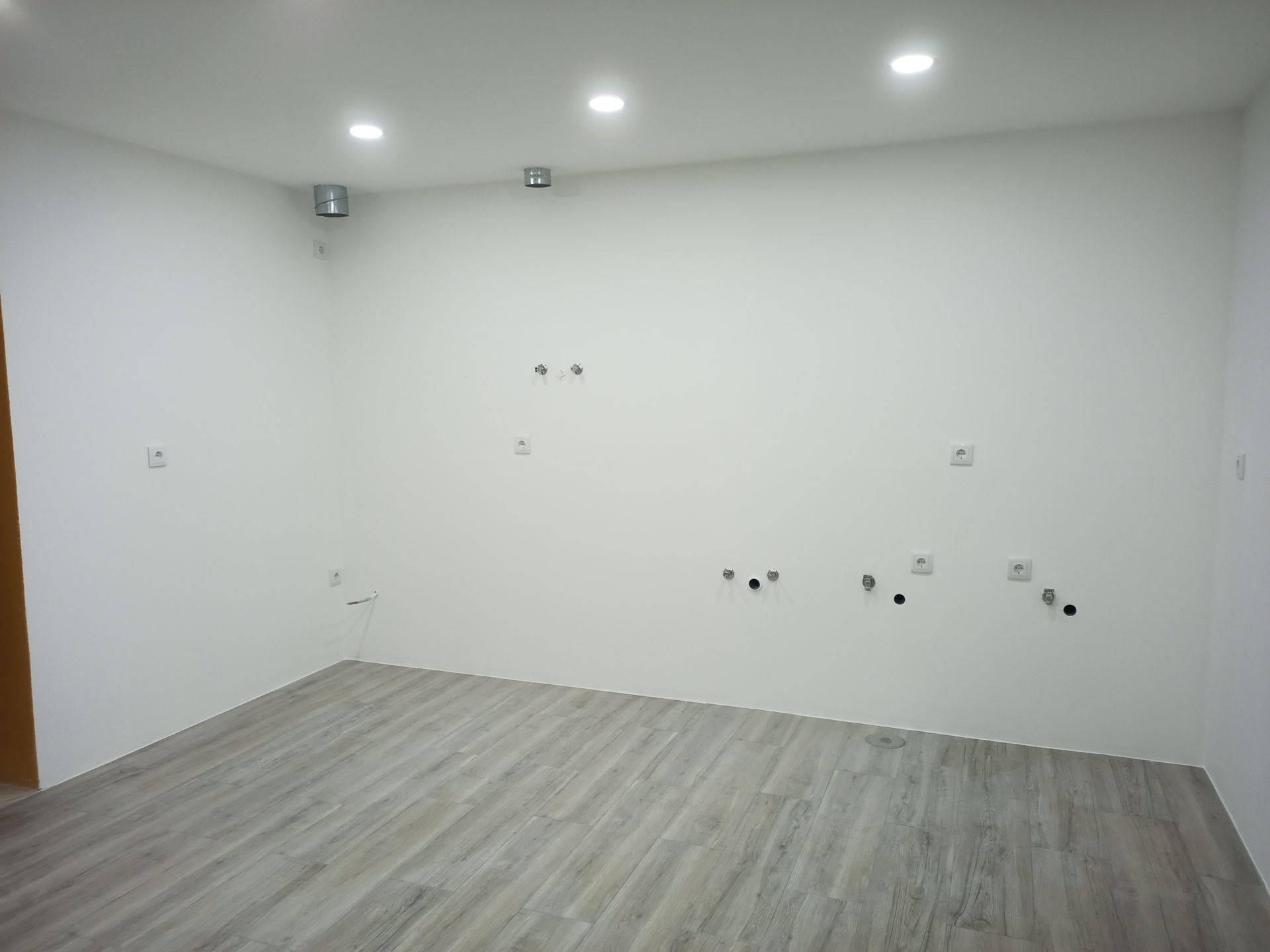 Sala vazia com piso imitando madeira em tom cinza claro e paredes brancas. Luminárias embutidas no teto e tomadas elétricas são visíveis.