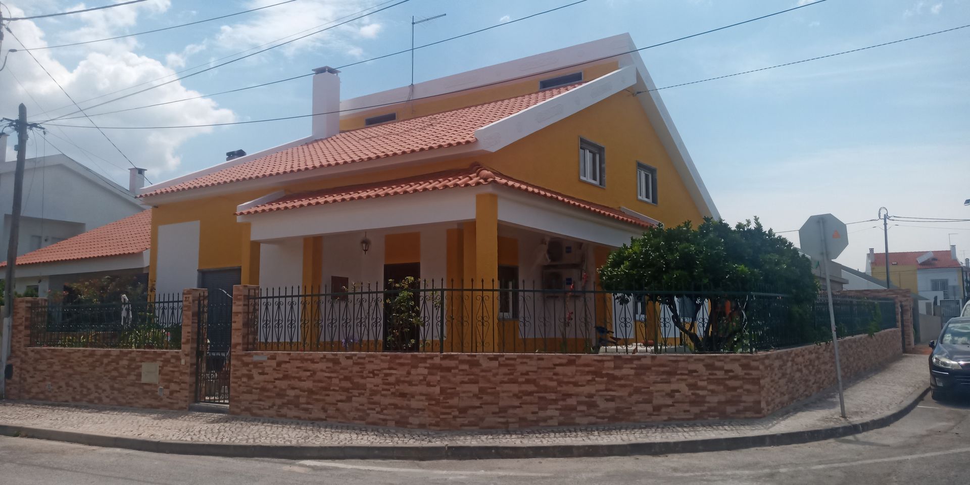 Casa amarela com telhado de telhas vermelhas, parede de tijolos e cerca de metal preta em um terreno de esquina.