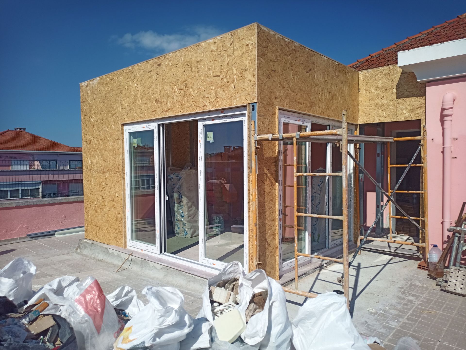 Canteiro de obras: paredes revestidas com OSB e portas de vidro deslizantes, andaimes e entulho em um telhado com um prédio rosa.