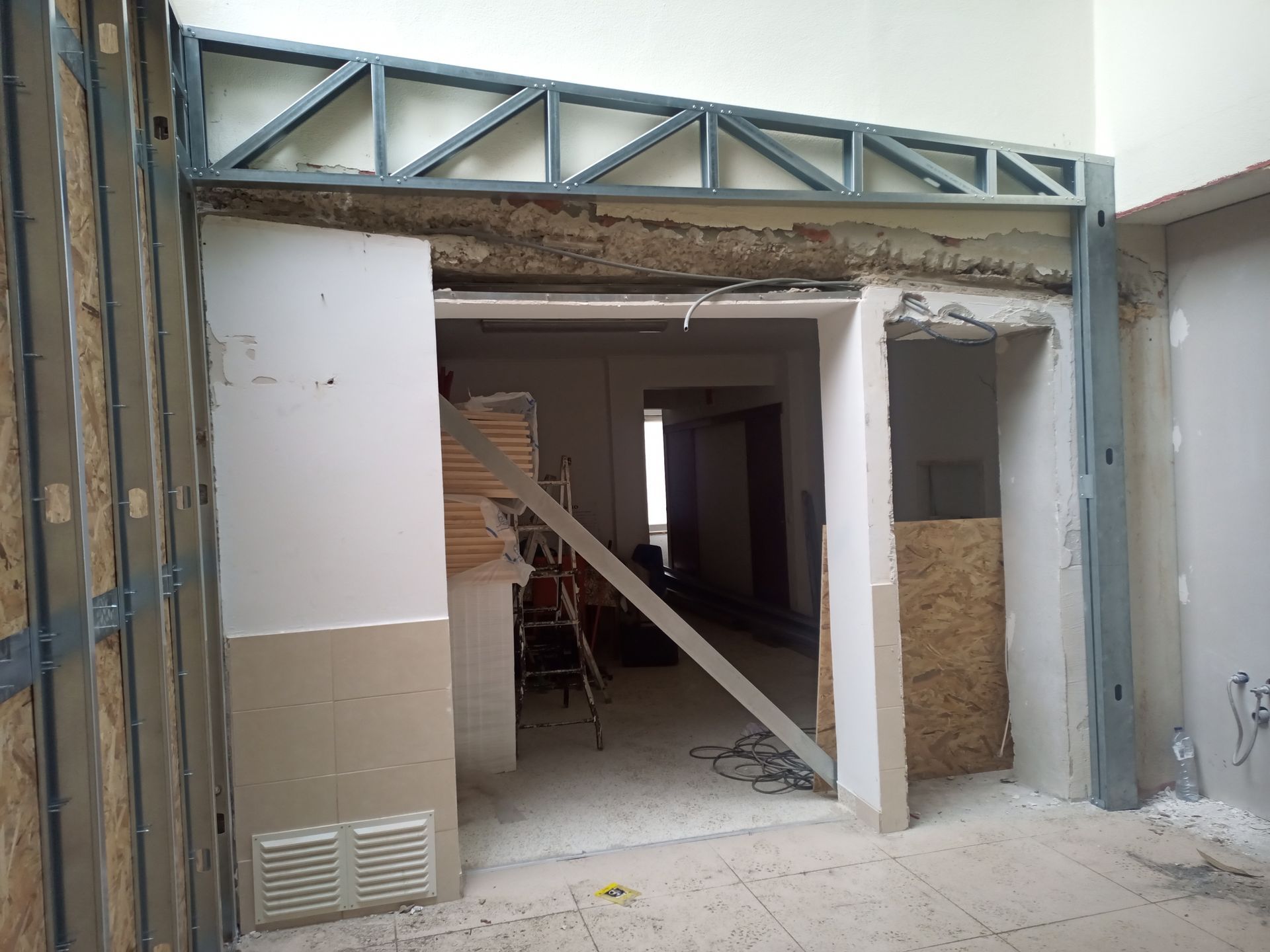 Interior em construção: porta de entrada com estrutura metálica, paredes inacabadas, compensado e teto parcial.