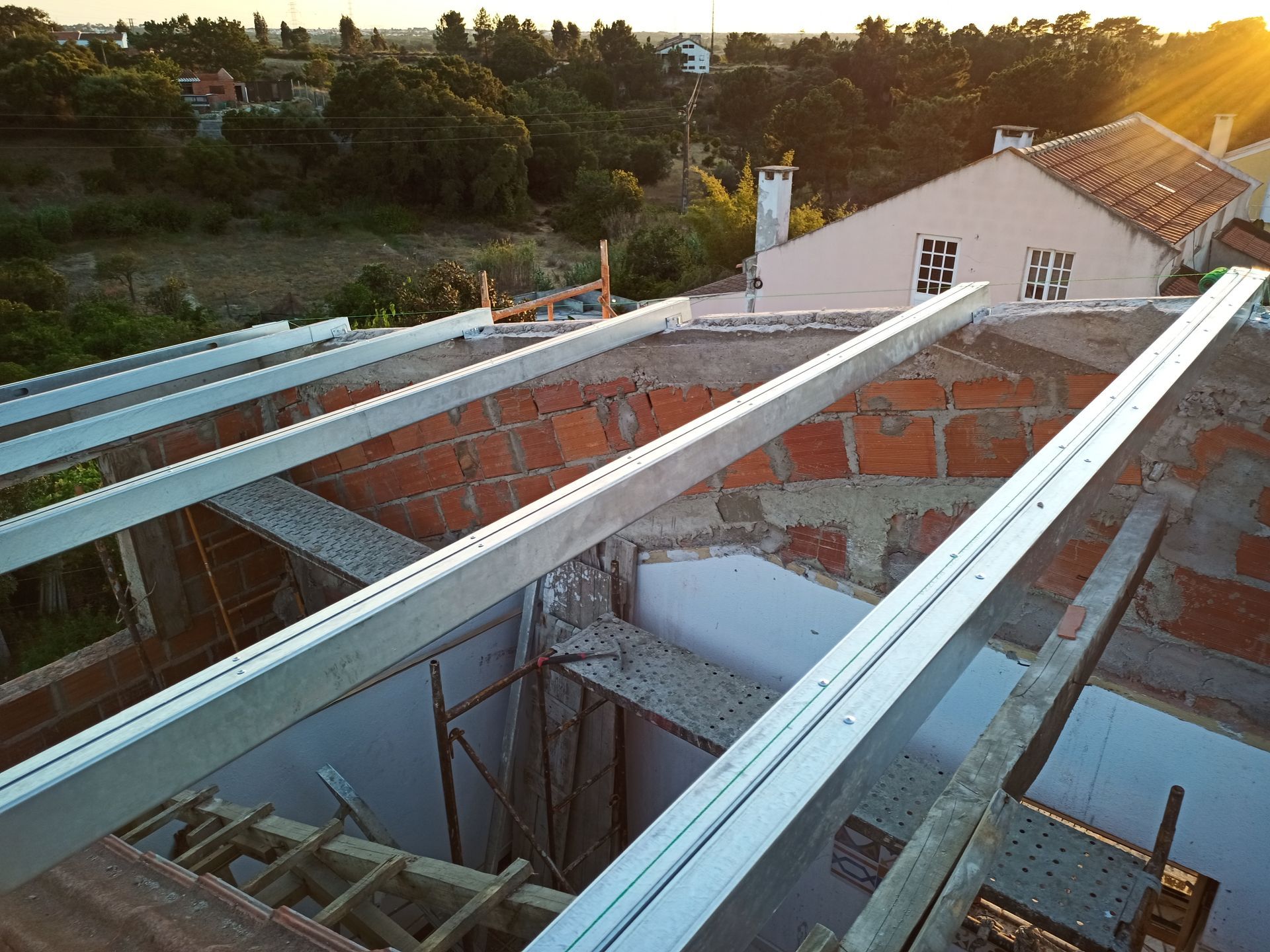 Canteiro de obras com vigas metálicas dispostas transversalmente sobre uma estrutura de tijolos; a luz do sol ilumina a cena.